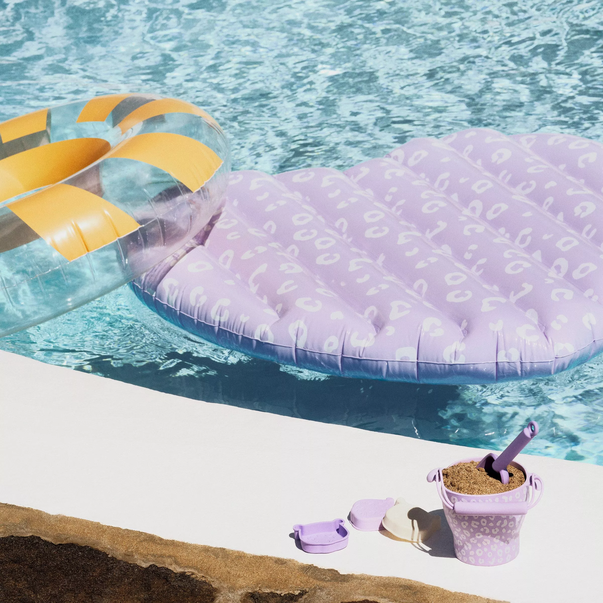 Inflatable pool floats, one purple with patterns, another striped, beside a pool with a sand-filled bucket.