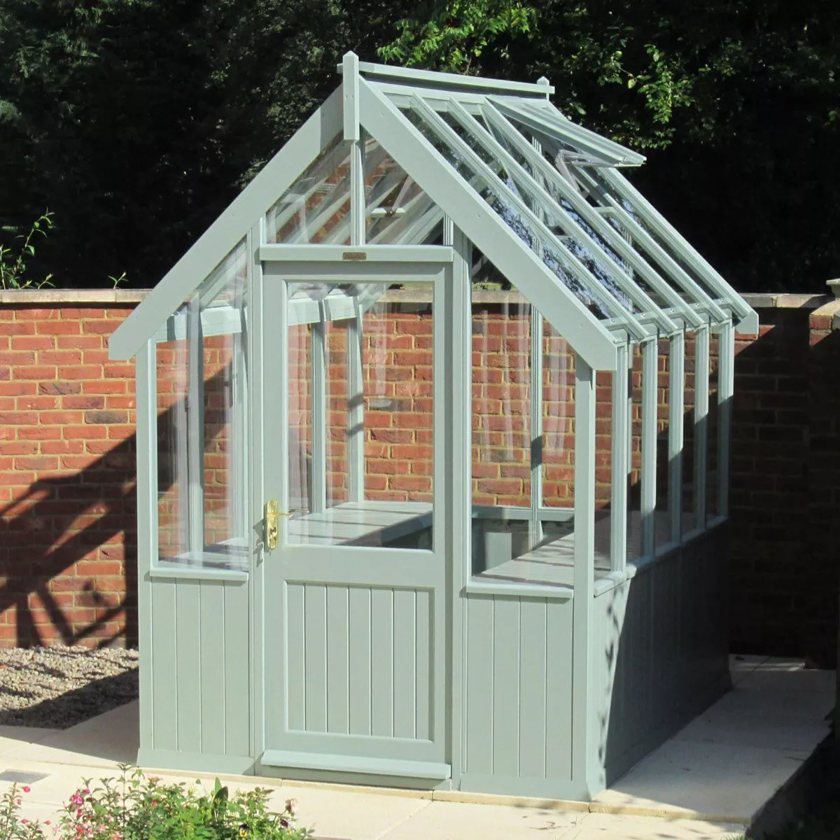 Light grey glass greenhouse with pitched roof and door, set on patio beside a red brick garden wall.