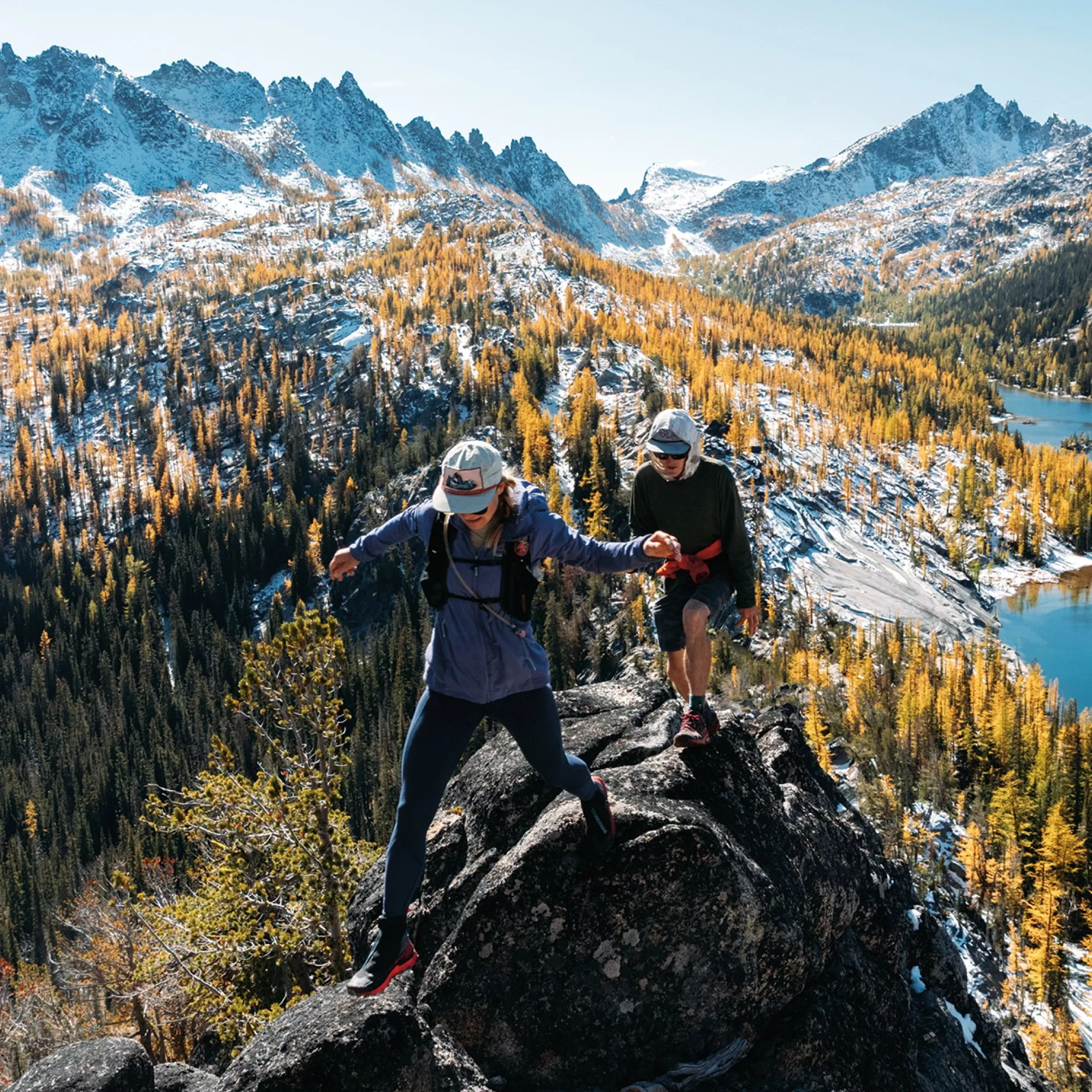 Two hikers scrambling on rocky ridge above alpine lake, autumn larch forest and snowy mountain peaks backdrop