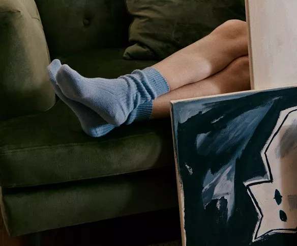 A person in blue socks lounges on a green sofa, with a dark abstract painting leaning nearby.