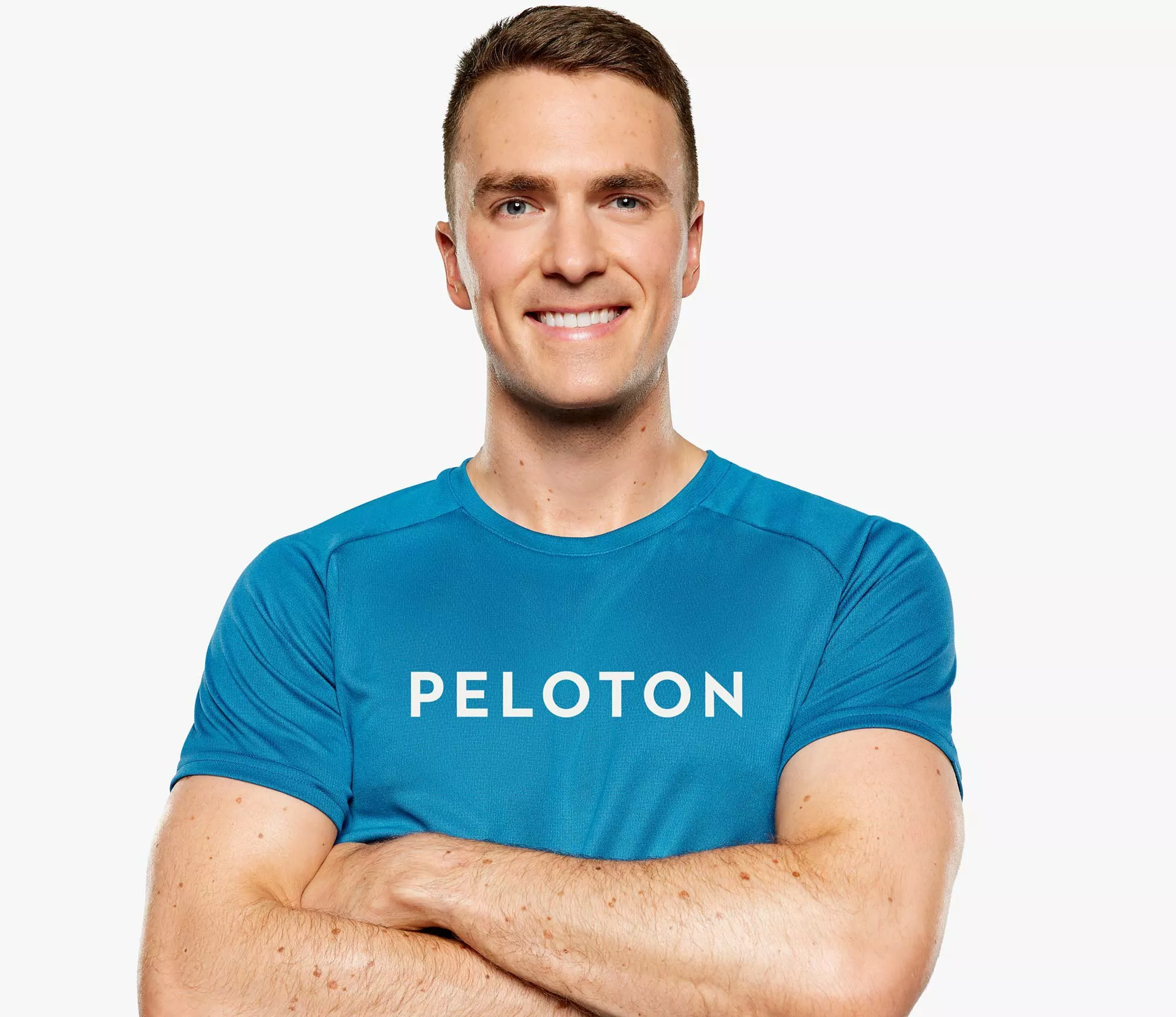 Smiling fitness instructor in blue Peloton T-shirt, arms folded, studio headshot on white background