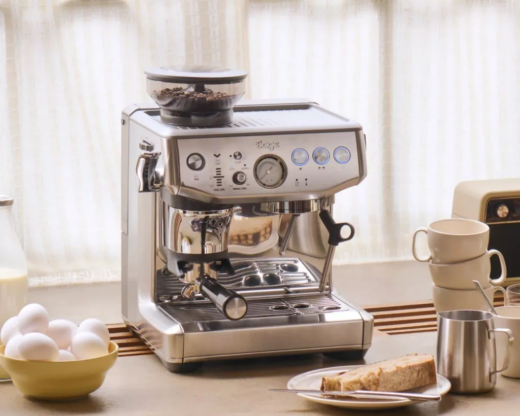 Stainless steel espresso machine with grinder on kitchen counter, beside cups, eggs, milk and toast in morning light
