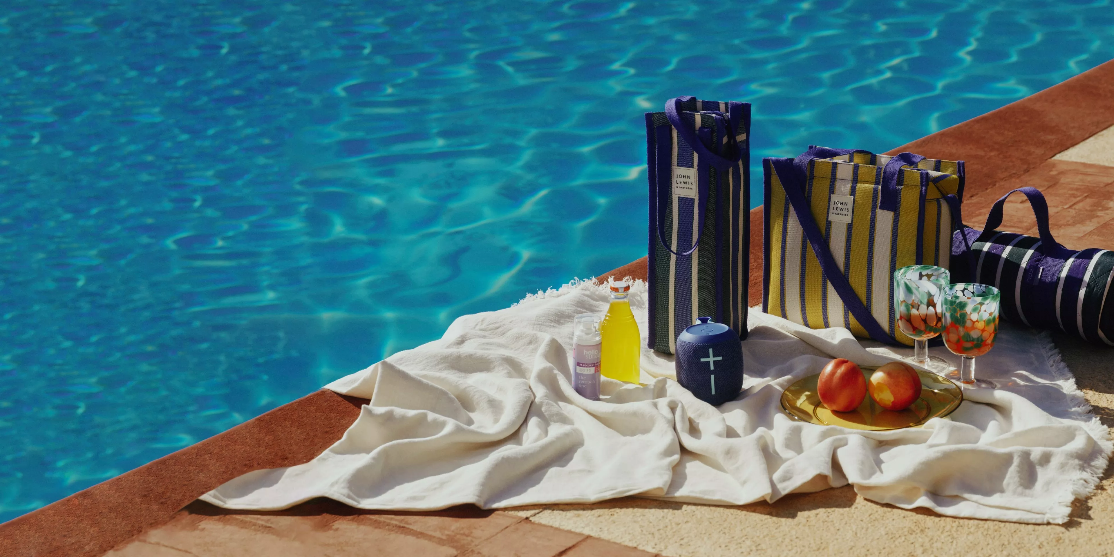 Poolside picnic setup with striped tote bags, blanket, drinks, fruit and glasses beside a sparkling blue pool