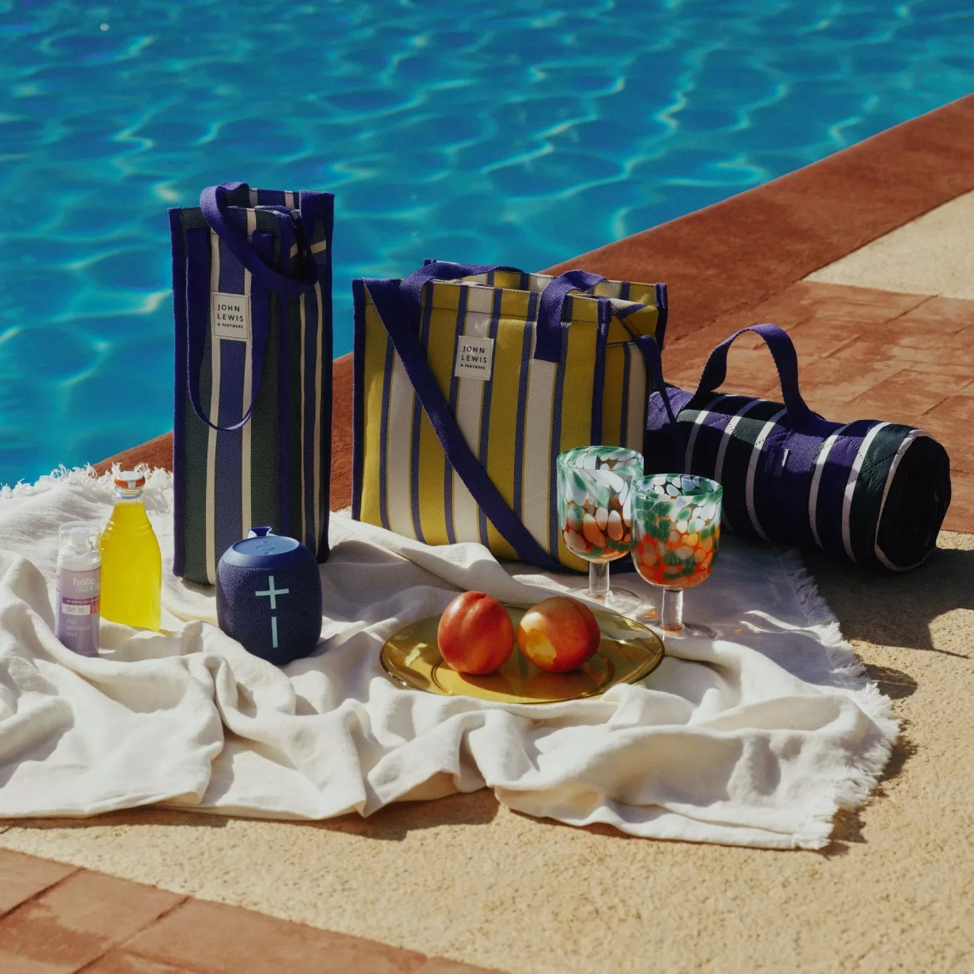 Poolside picnic set with striped tote bags, blanket, fruit, drinks, suncream and portable speaker by the pool