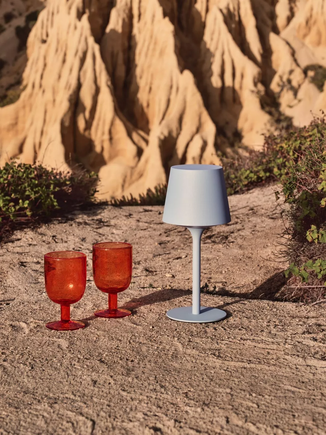 Modern grey cordless table lamp with two amber glass tumblers on sandy ground, rocky cliff backdrop outdoors