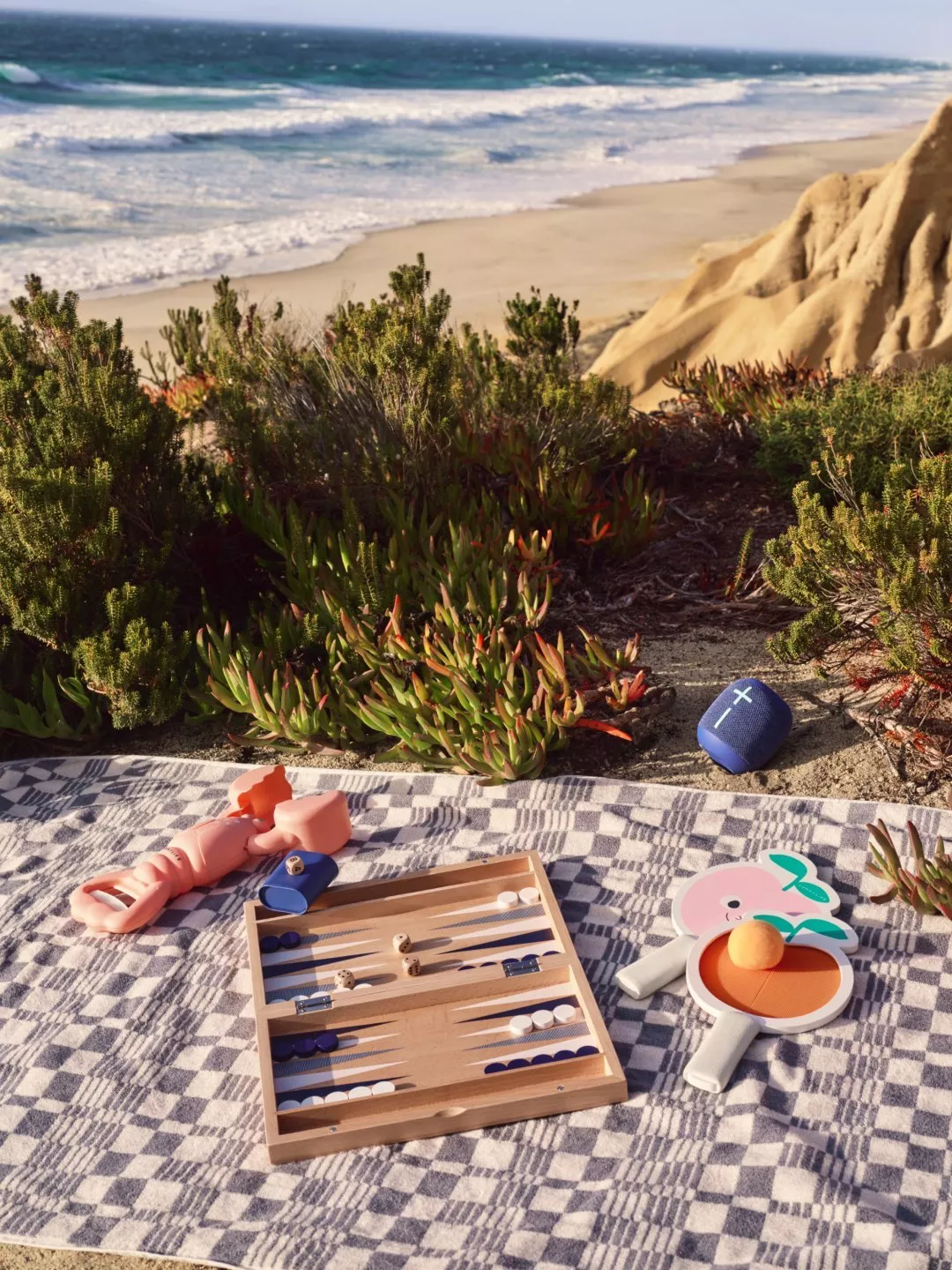 Coastal beach picnic blanket with backgammon set, paddles, toy and portable speaker beside sand dunes and surf.