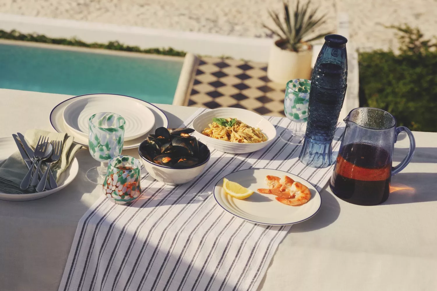 Outdoor seafood lunch table by pool with prawns, mussels, pasta, wine jug and blue glass bottle in sun