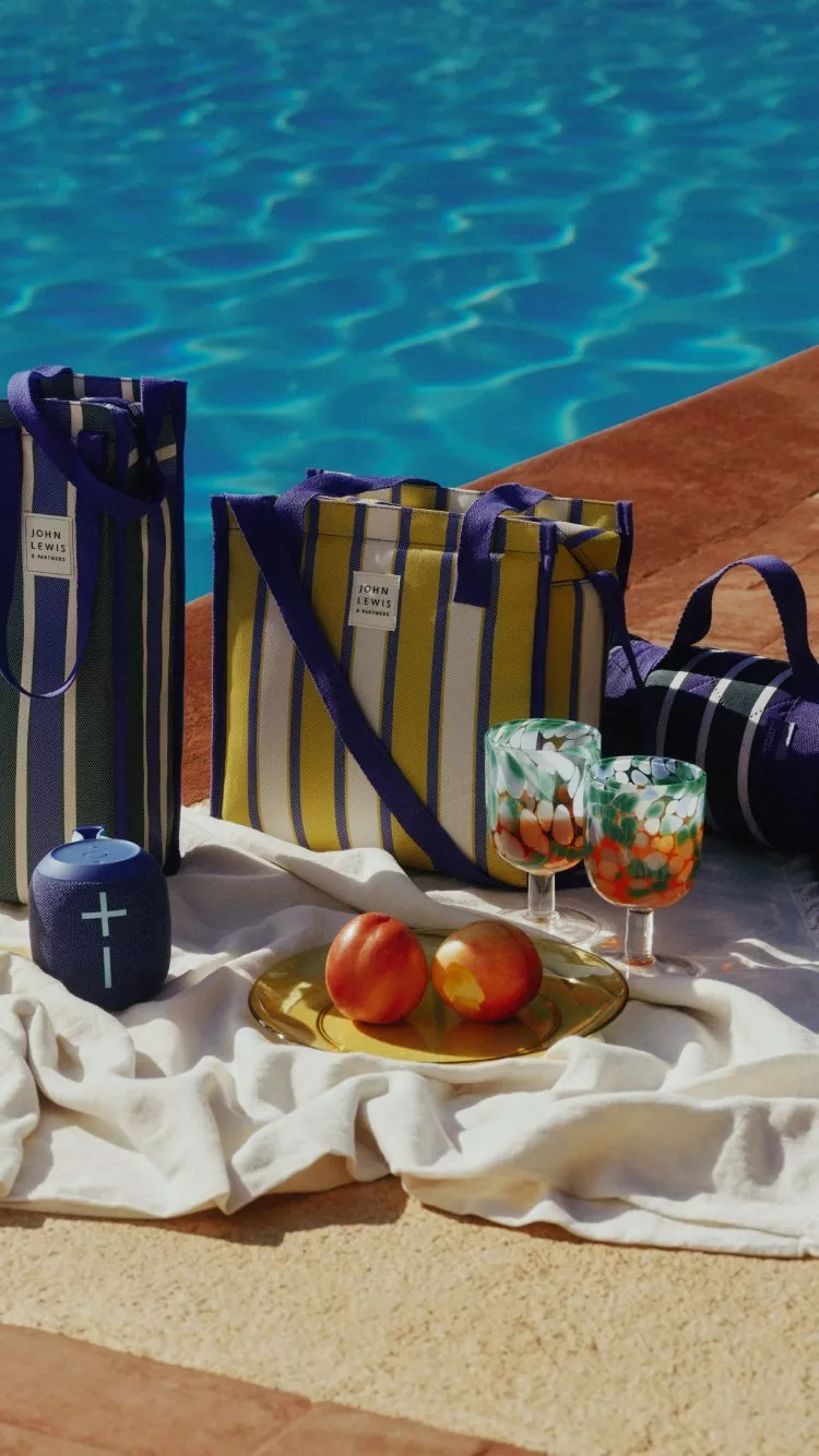 Poolside picnic setup with striped tote bags, patterned glasses, peaches on tray and portable speaker by water