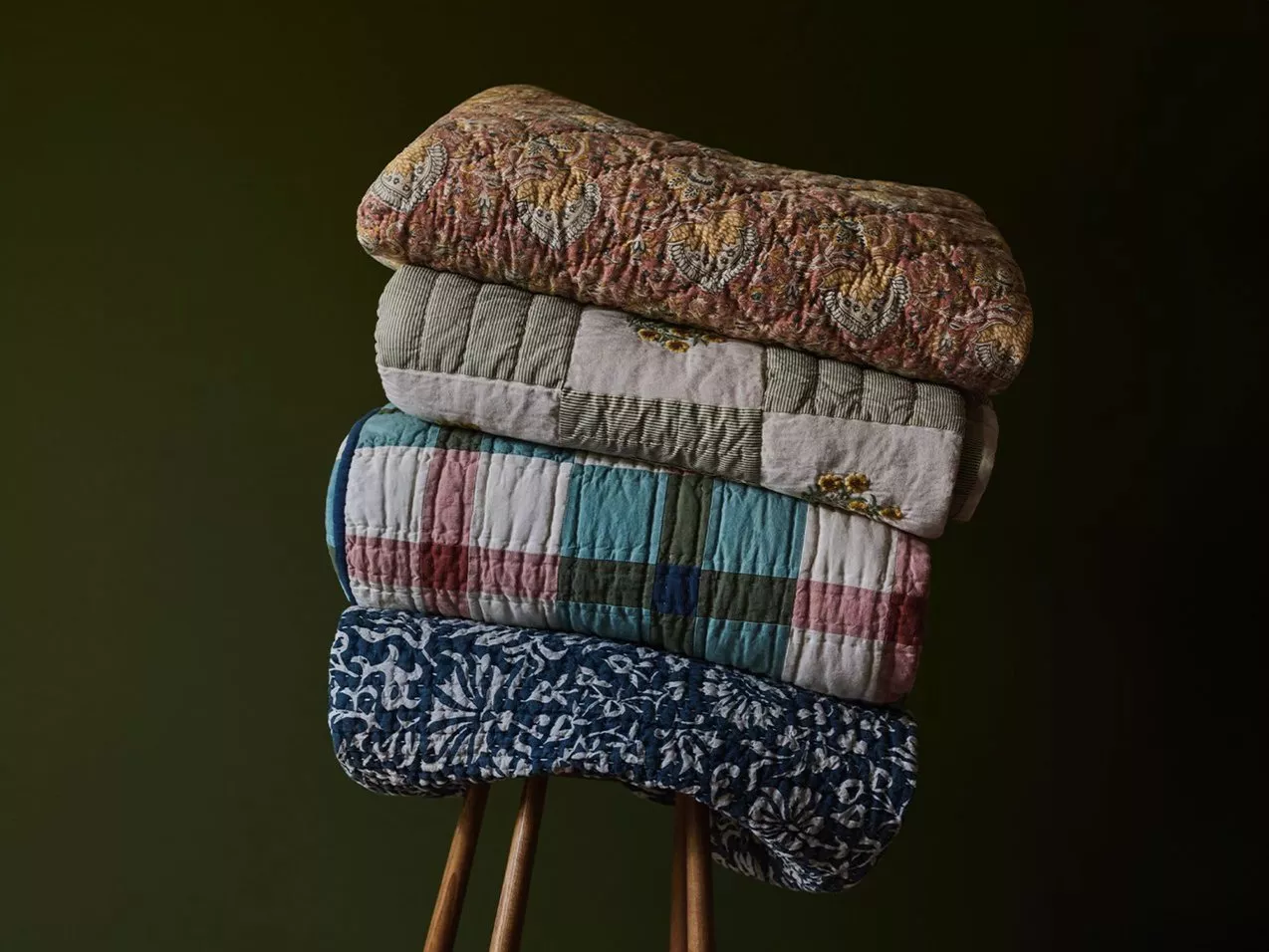4 quilted throws stacked on-top of one another on a wooden stool with a green background