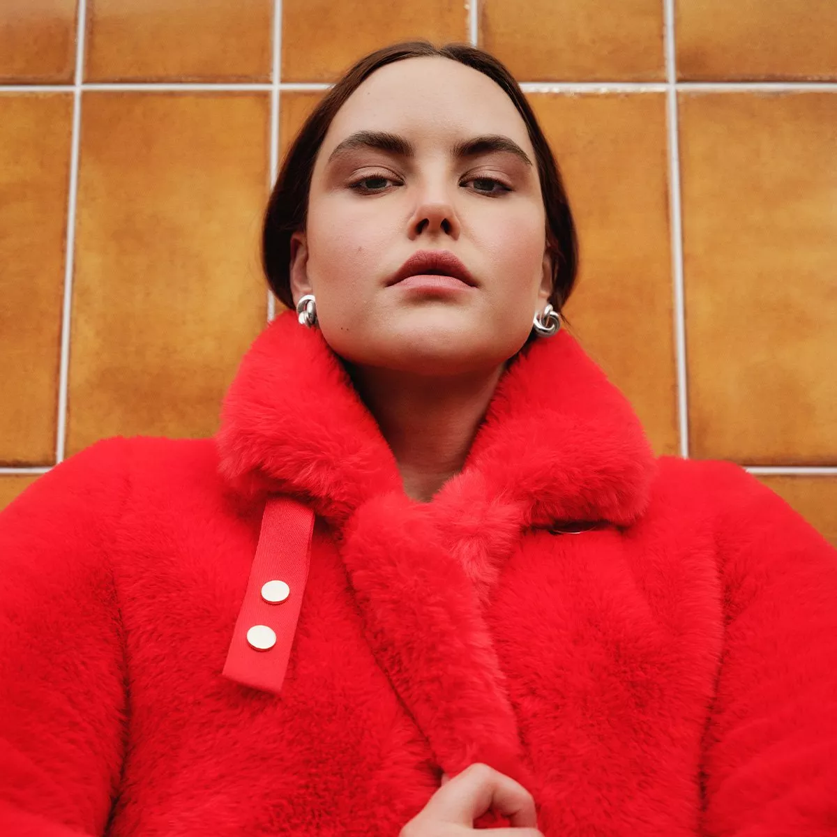 Woman in a bold red fur coat poses confidently against a brown tiled wall, wearing silver earrings.