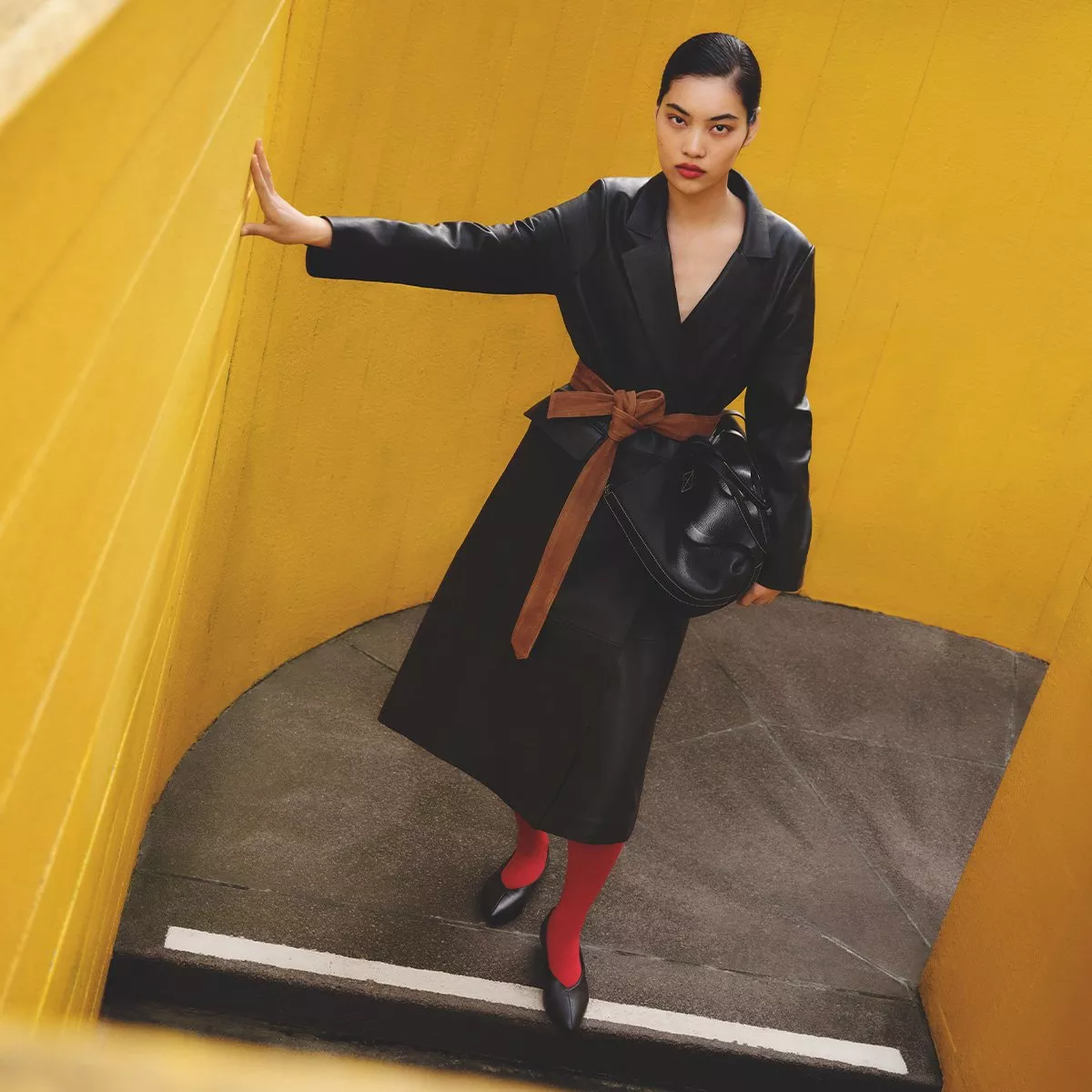 Stylish person in black coat, red tights and brown belt, leaning against bright yellow wall outdoors.