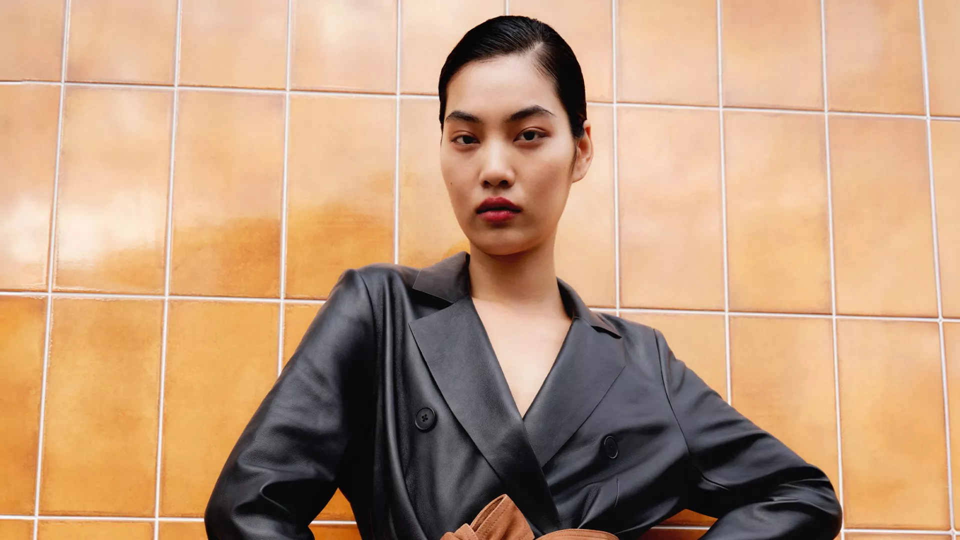 Model in black leather coat with brown belt poses confidently against orange tiled wall.