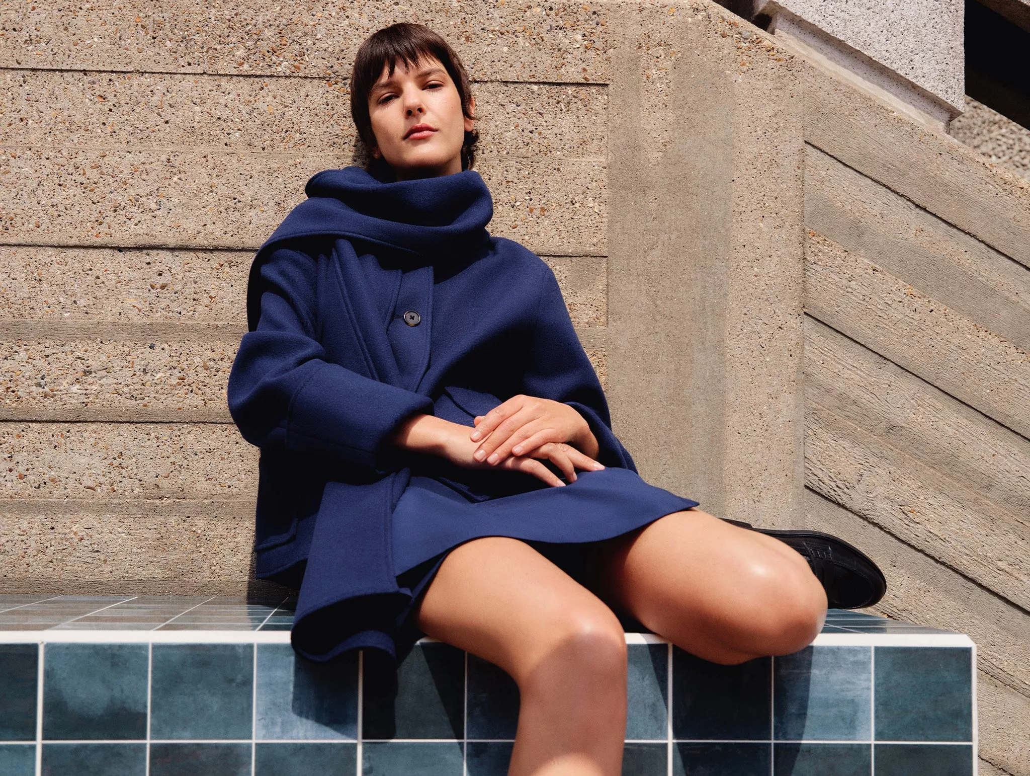 A person in a stylish blue coat sits on a tiled surface against a textured concrete backdrop, exuding confidence.