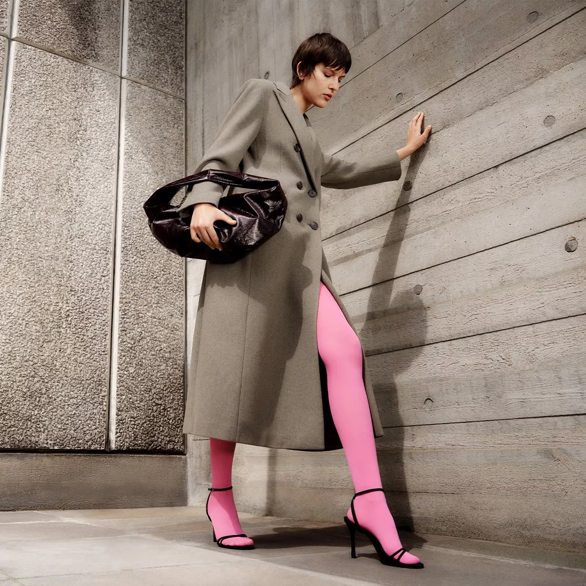 Fashionable person in a long coat, pink leggings, and black heels, carrying a large handbag, leans on a wall.