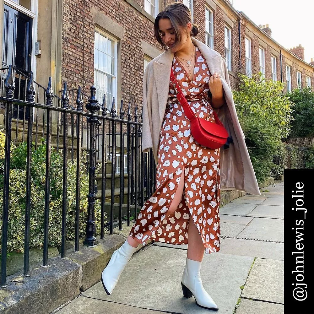 Stylish woman in a printed dress with a beige coat, red crossbody bag, and white boots on a cobbled street.