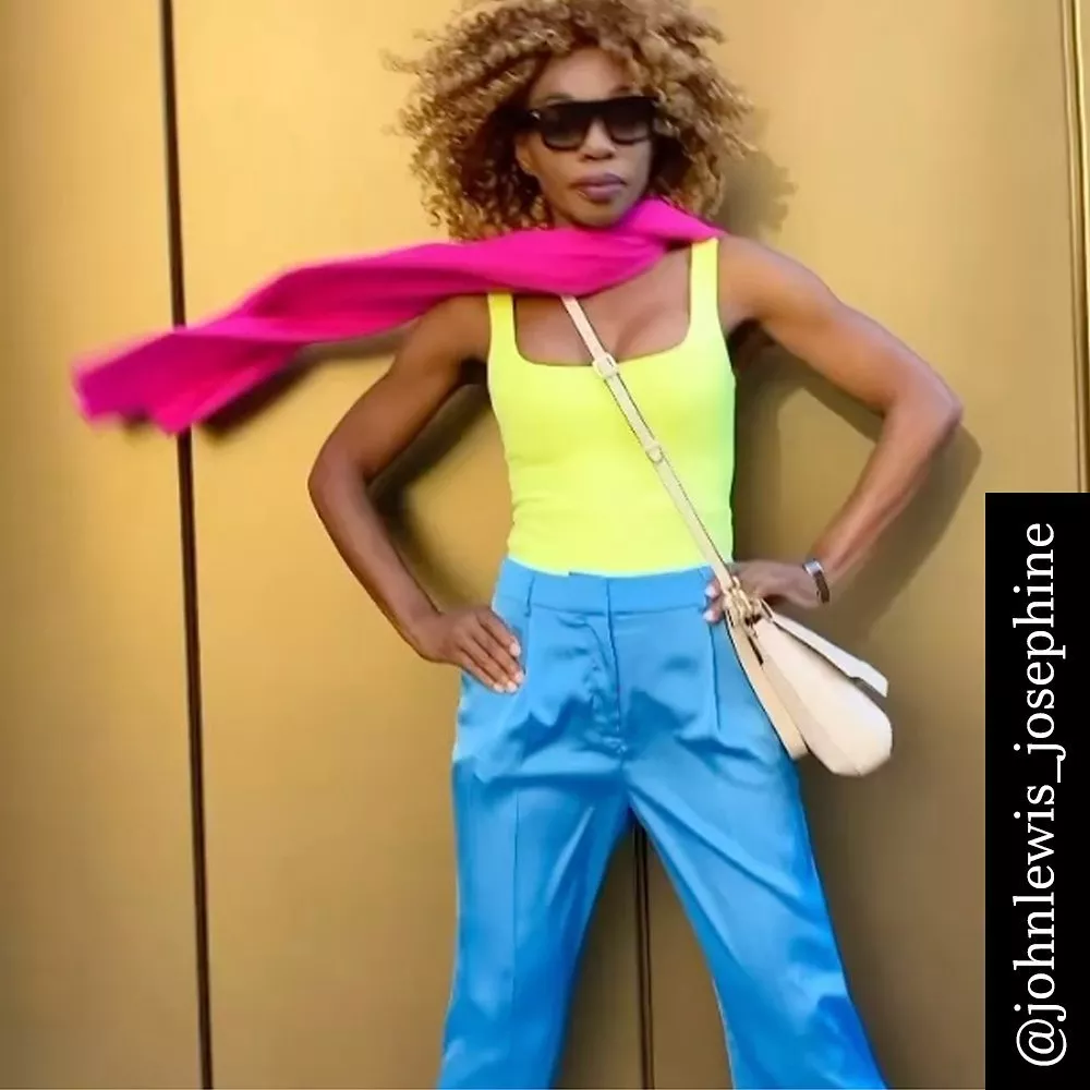 Fashionable person in bright yellow top, blue trousers, pink scarf, and sunglasses against a gold background.