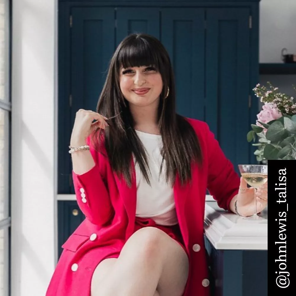 A woman in a pink blazer sits elegantly, holding a drink. Flowers decorate the table near her.