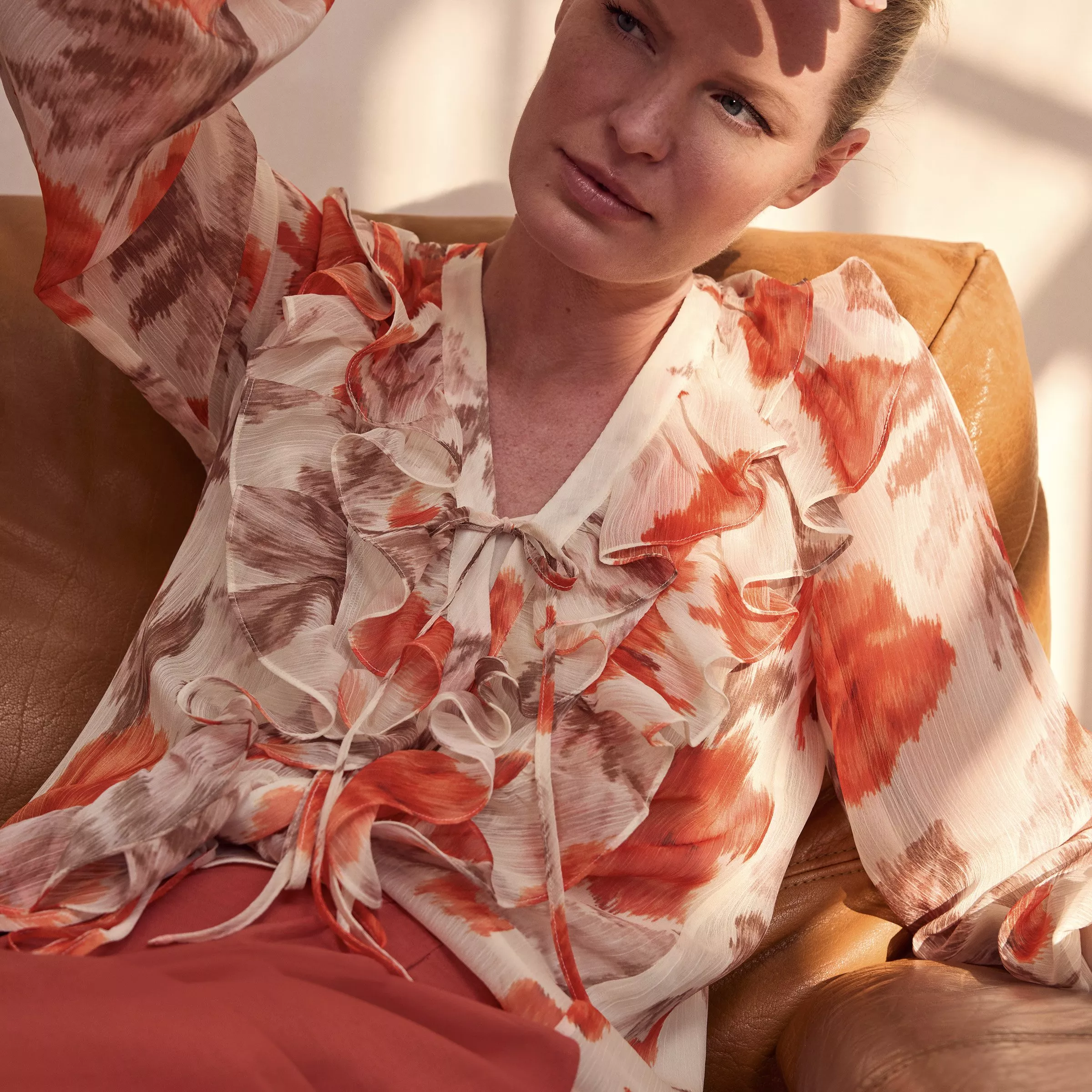 Woman lounging on tan leather sofa wearing ruffled floral chiffon blouse and coral trousers in warm sunlight