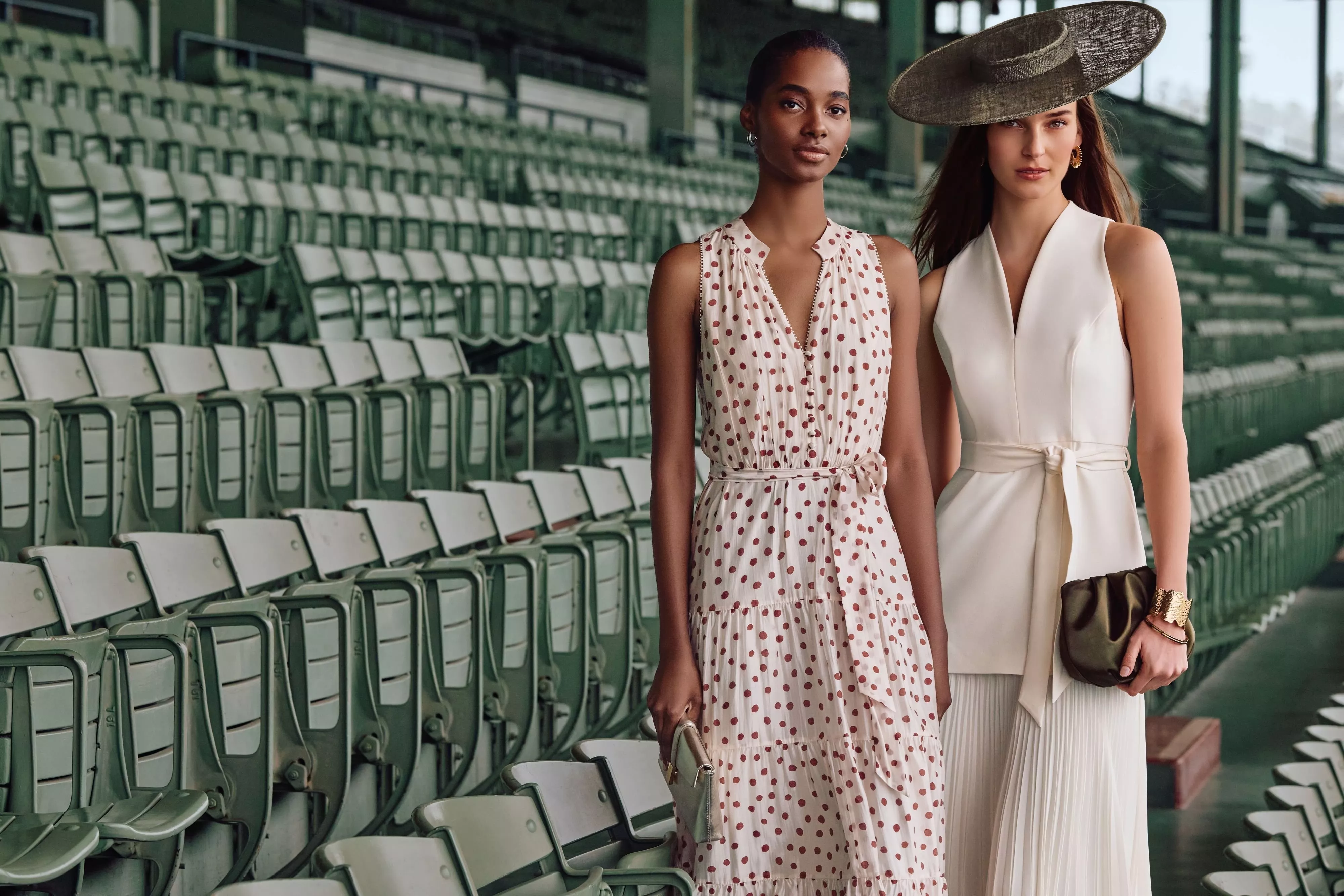 Two stylish women in elegant dresses and hats posing in empty grandstand seats at a racecourse fashion event
