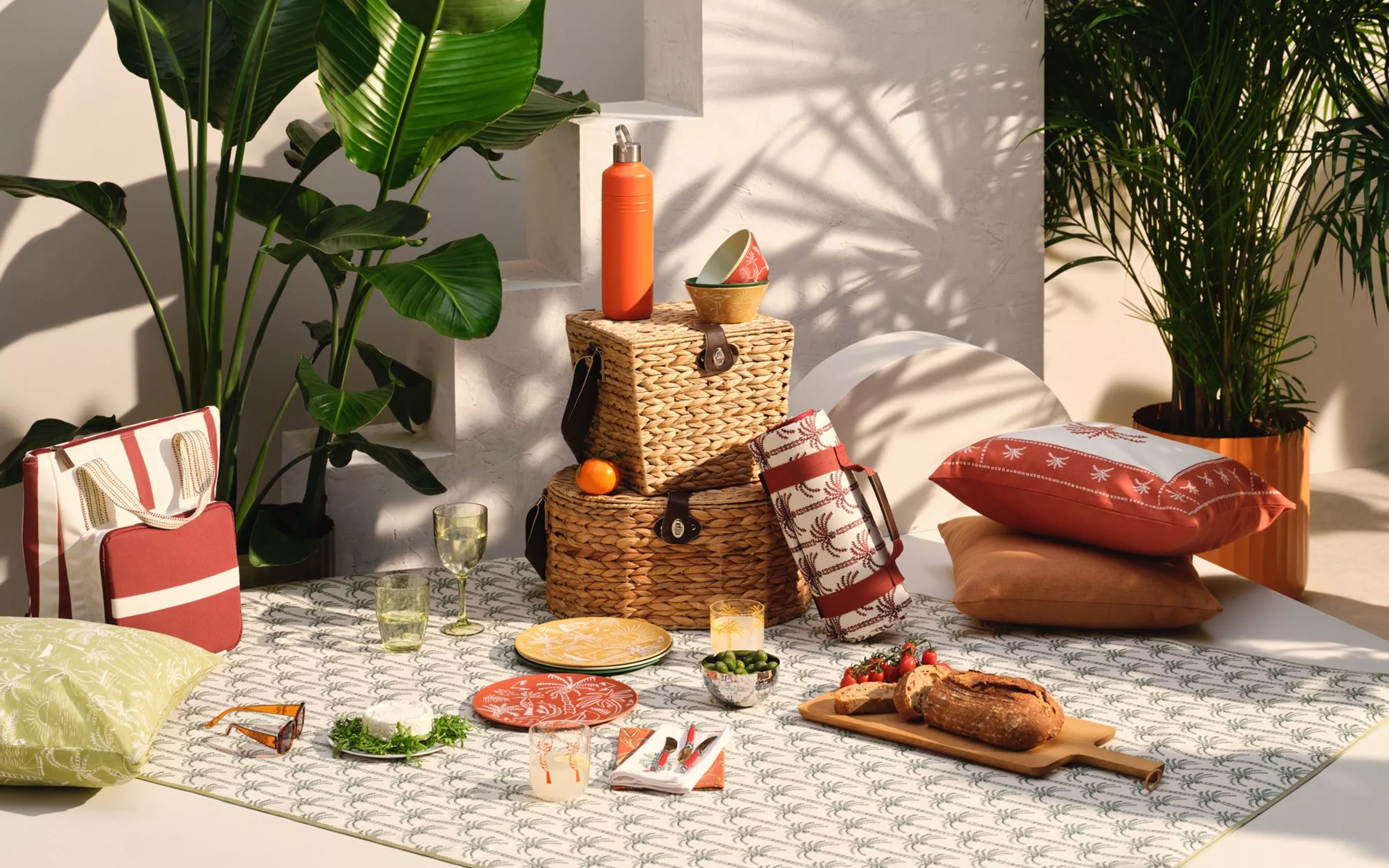 Sunlit picnic setup with patterned blanket, woven baskets, cushions, bread board, glasses and plants indoors