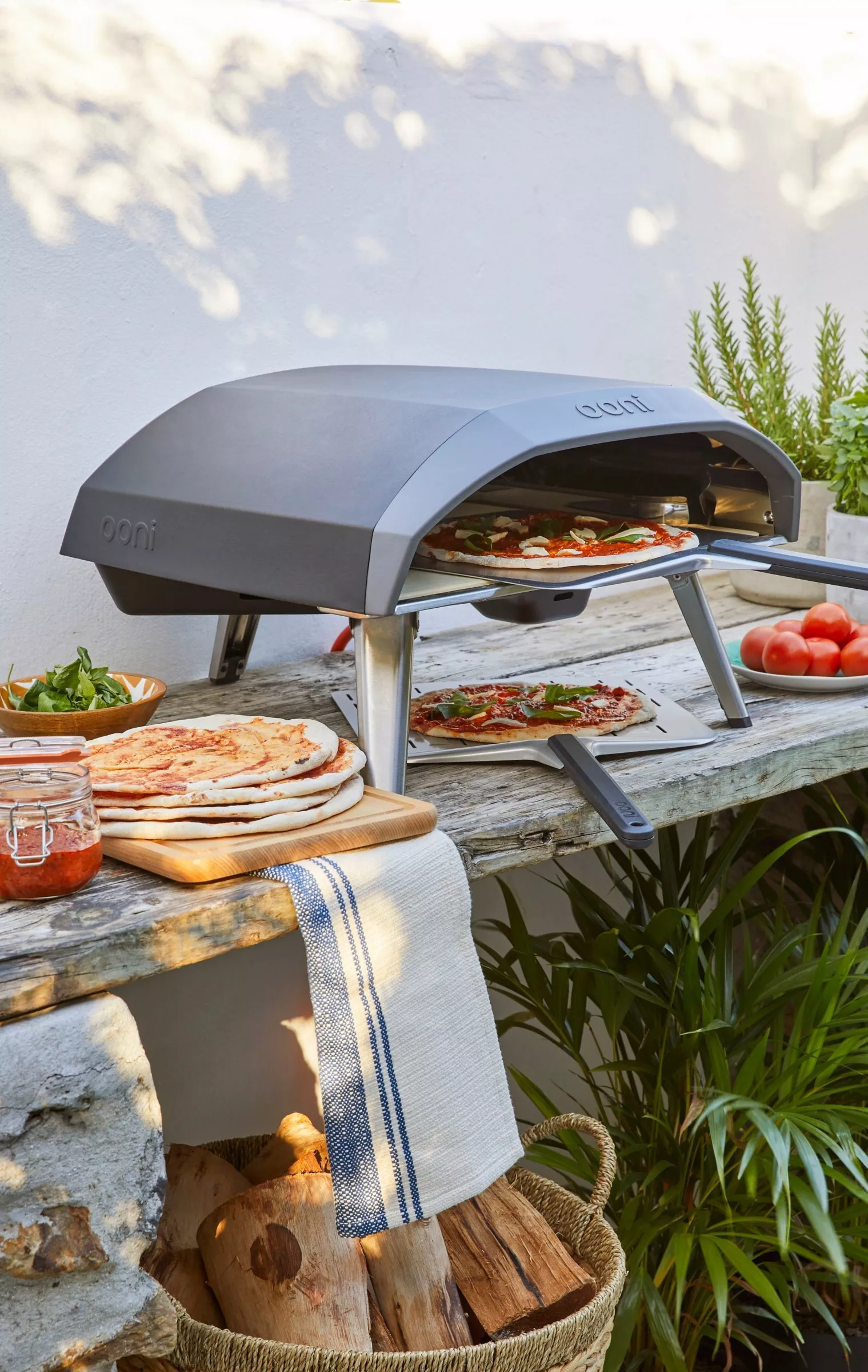 Outdoor pizza oven baking a fresh Margherita pizza on a wooden table with dough bases, herbs and tomatoes