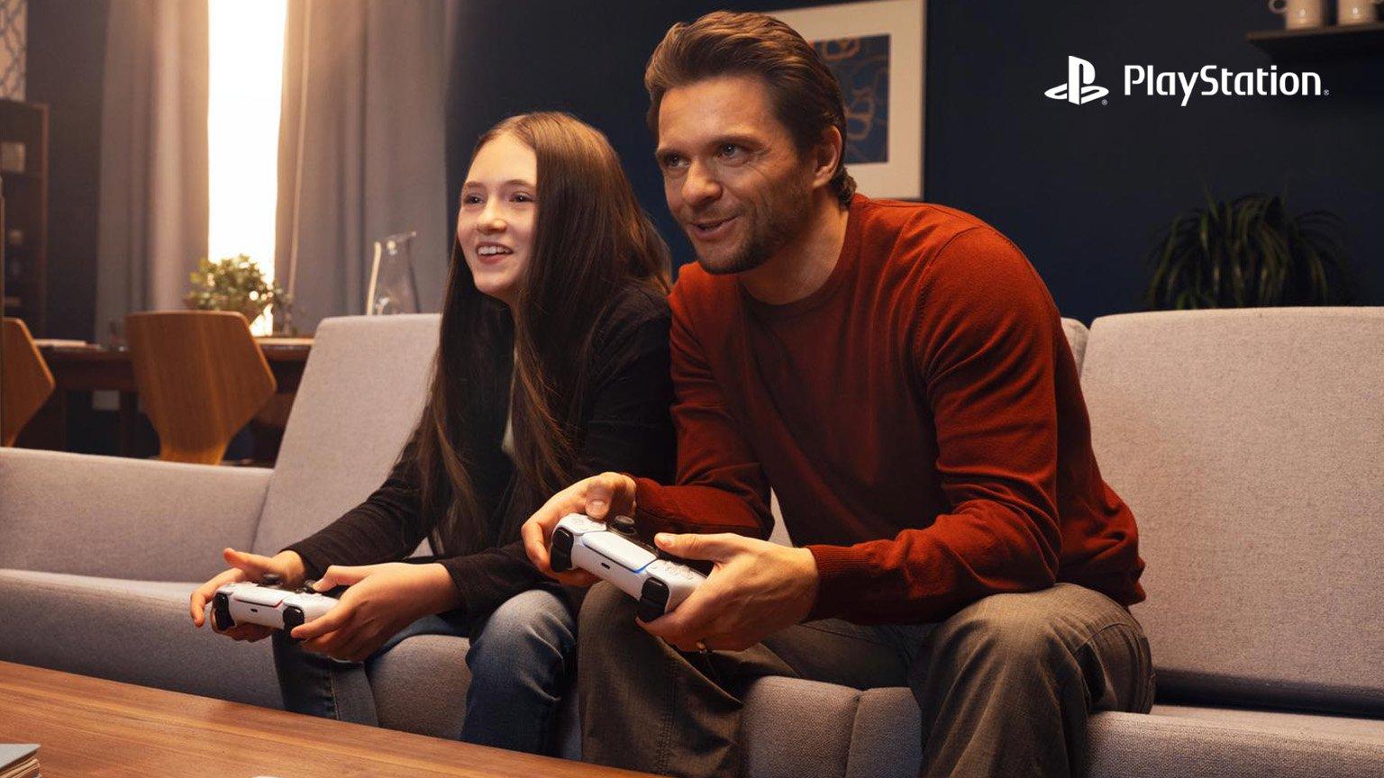 A man and a young woman sit on a gray couch, playing video games together in a cozy, warmly lit living room. A wooden coffee table with coasters and a notebook is in front of them. The background features a dining area and modern decor