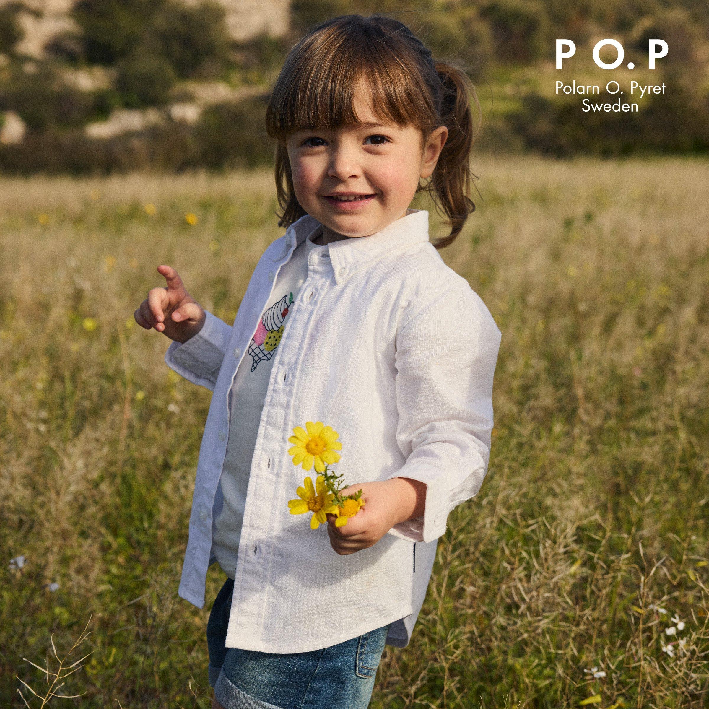 A young child stands in a field holding yellow flowers, smiling and wearing a white shirt over a printed t-shirt and denim shorts