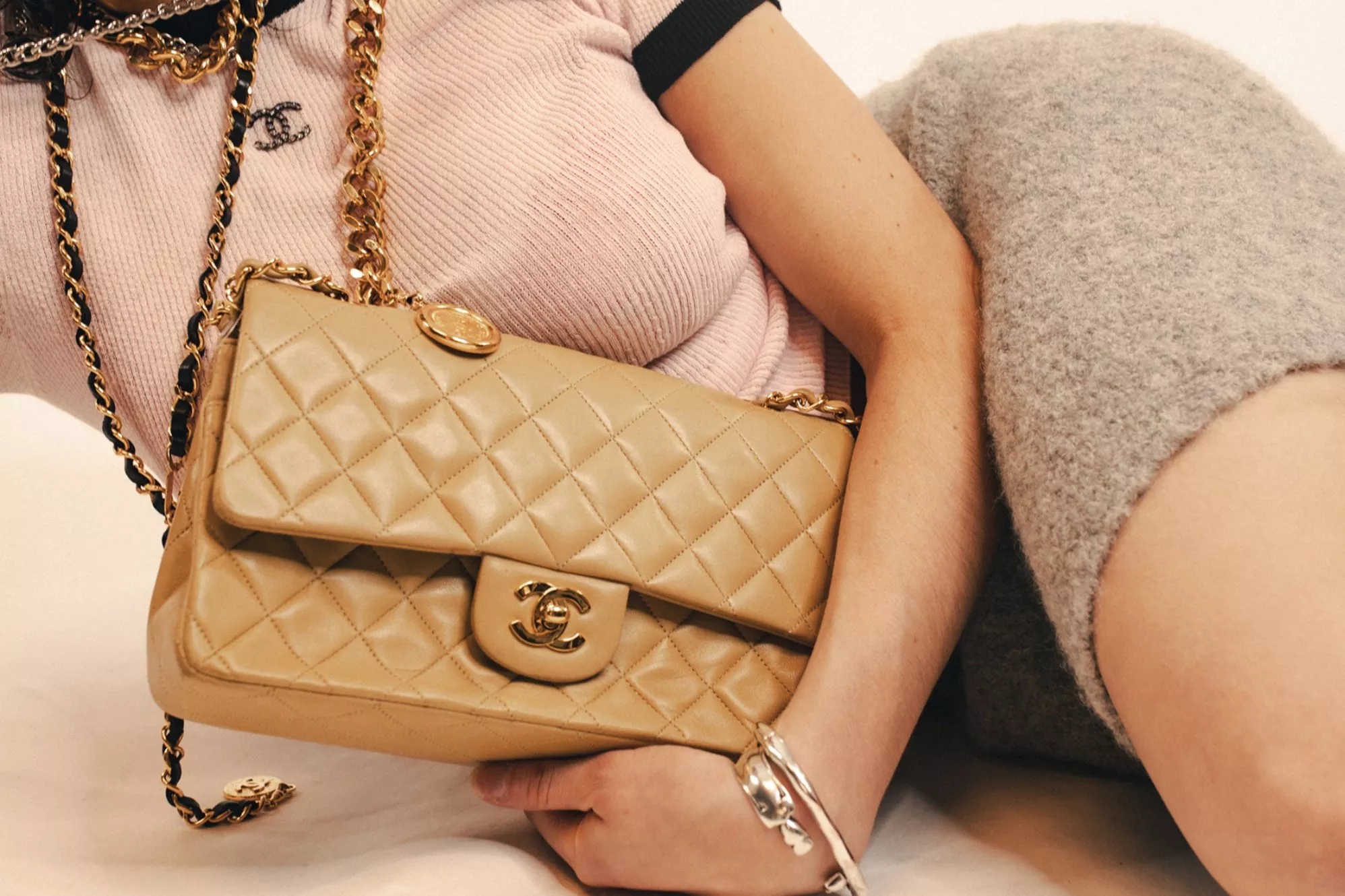 A person holds a stylish beige quilted handbag with gold chain, wearing a pink top and grey textured skirt.