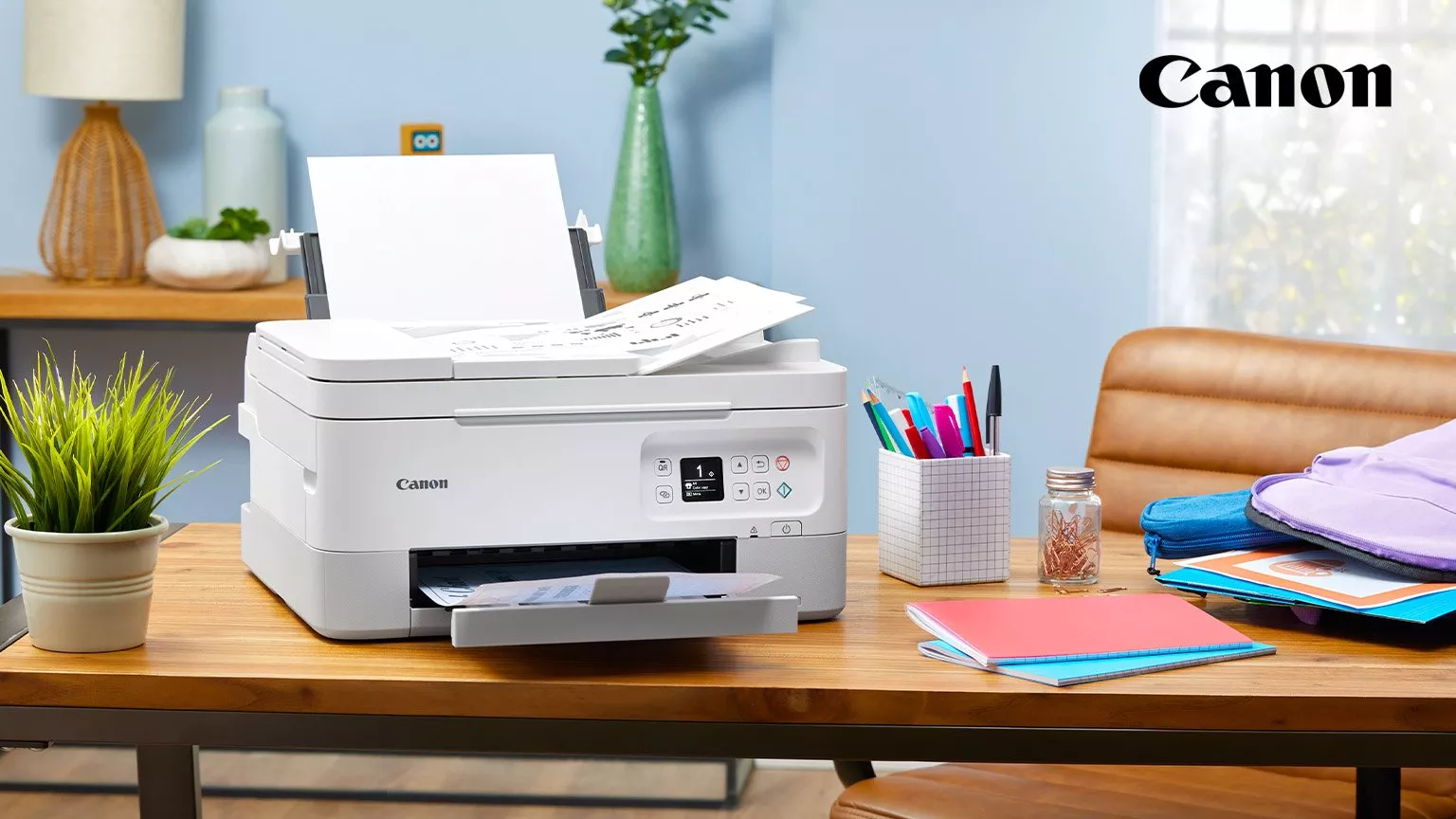 A Canon printer on a wooden desk, surrounded by stationery, notebooks, and a potted plant, in a home office.