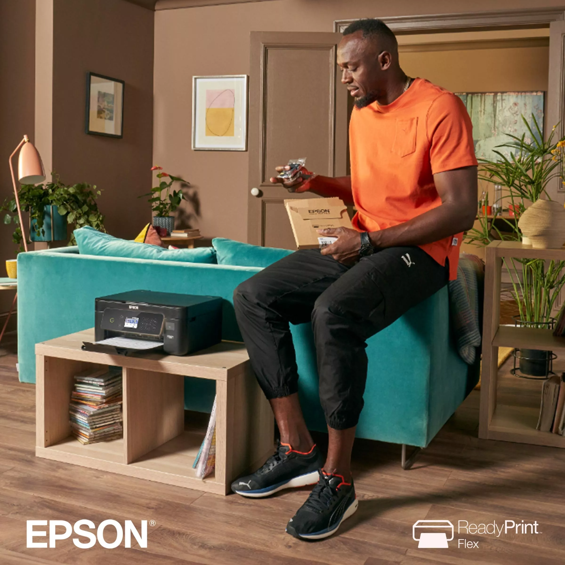 Man in orange shirt and black trousers sits on a sofa holding Epson printer ink, next to an Epson printer.
