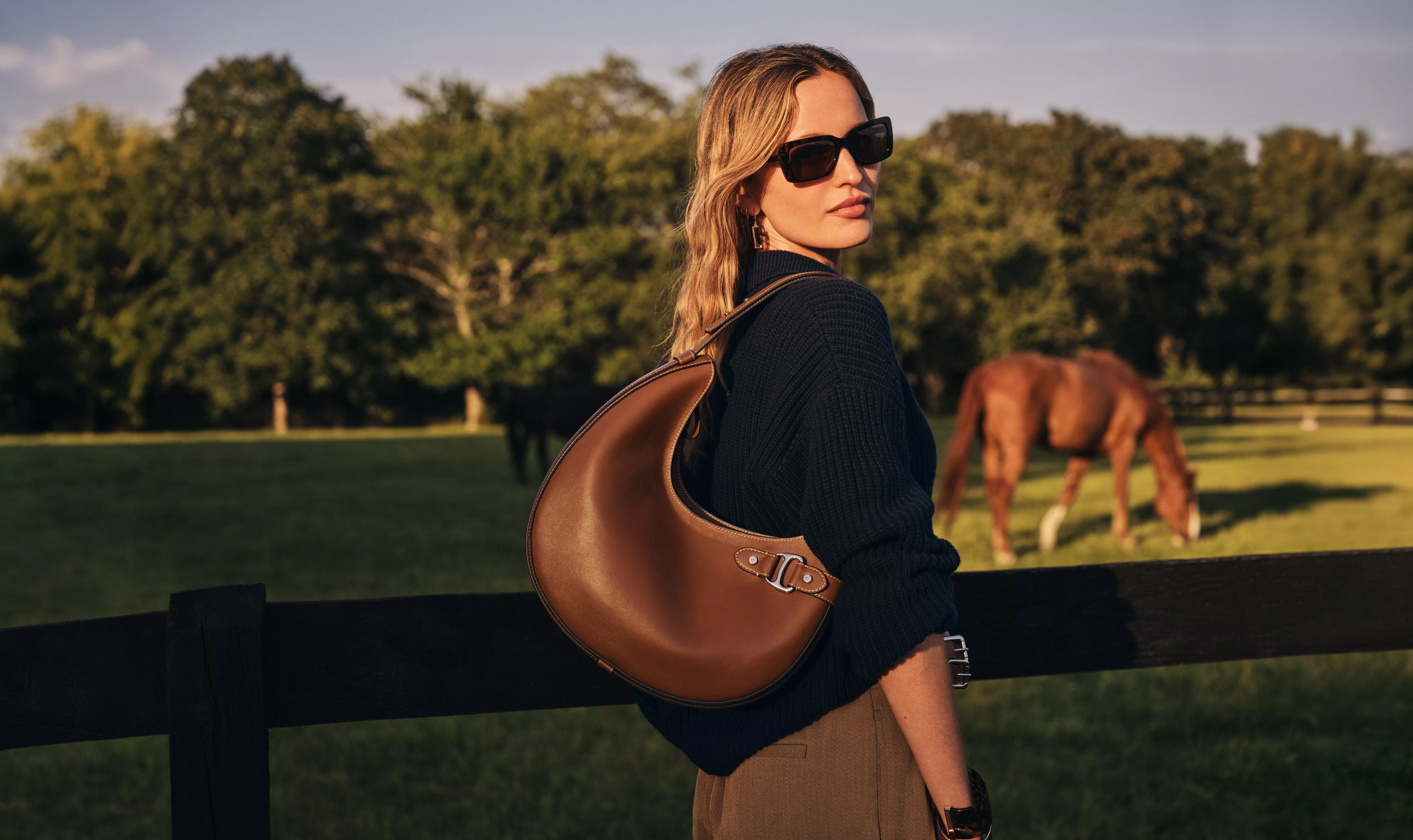 a woman standing in front of a horse field wearing dark shades, a black knitted jumper, and a brown leather handbag over one shoulder, all from Lauren Ralph Lauren