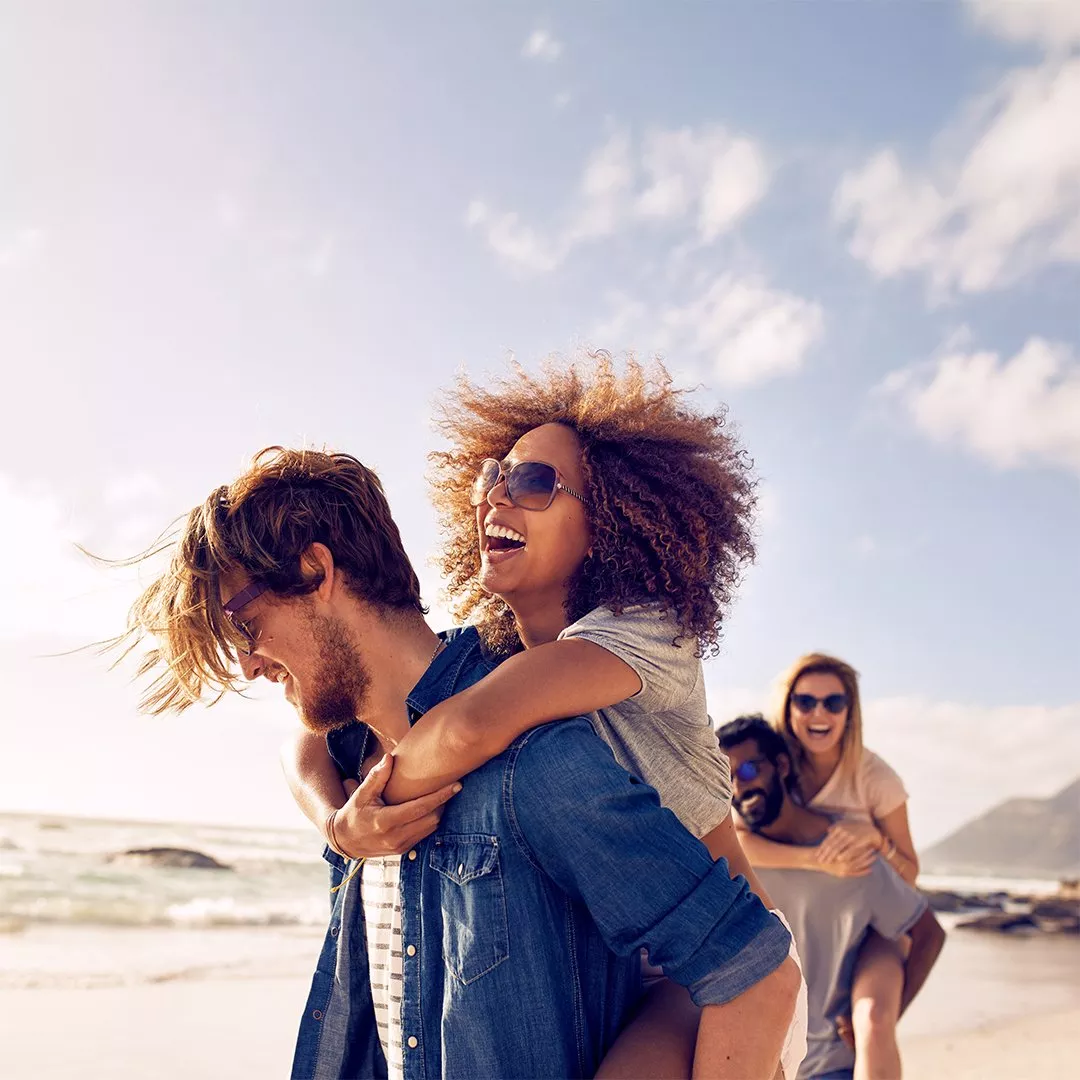 A joyful group of friends enjoying piggyback rides on a sunny beach, with clear skies and ocean waves in the background.