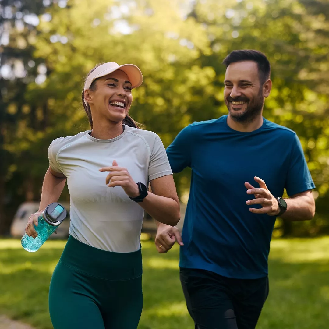 Two people jogging outdoors in athletic gear, surrounded by lush green trees and natural sunlight.