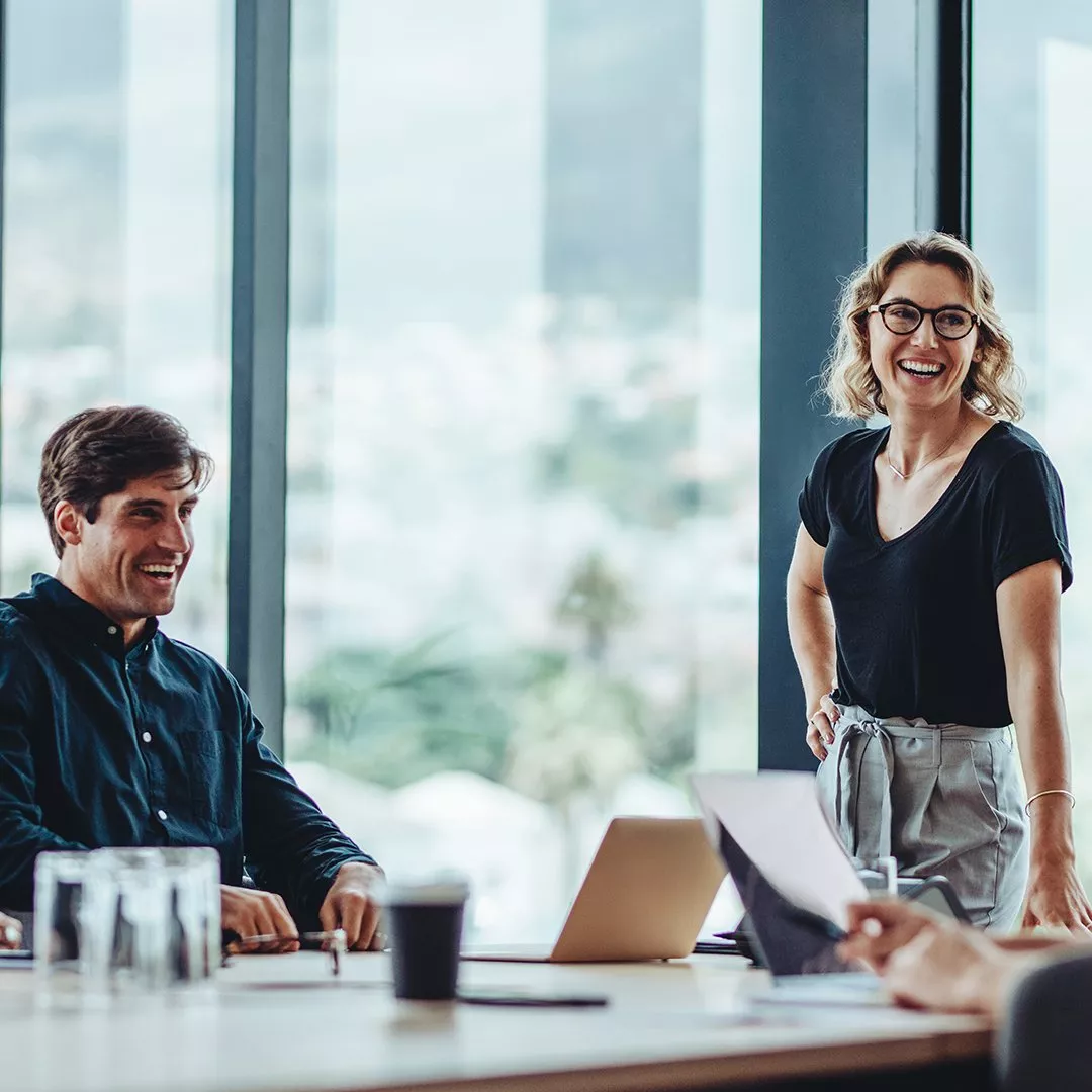 Business meeting in modern office with large windows. People are discussing around a table with laptops and documents.