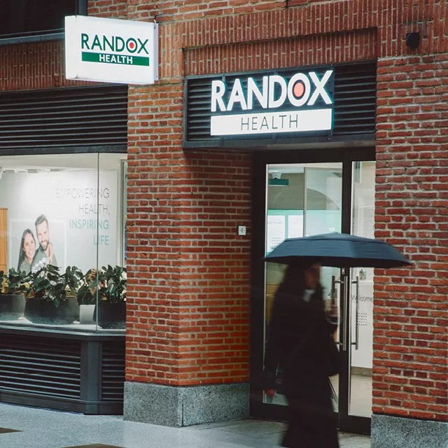 Randox Health clinic entrance on brick building with illuminated sign and person walking with umbrella outside