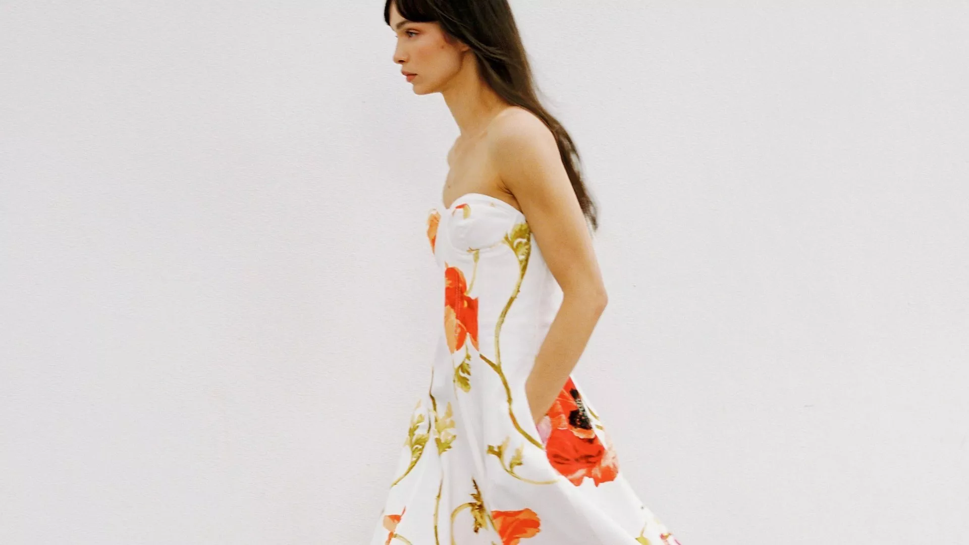 Woman in a strapless white dress with bold floral print walking against a plain white background. Elegant style.