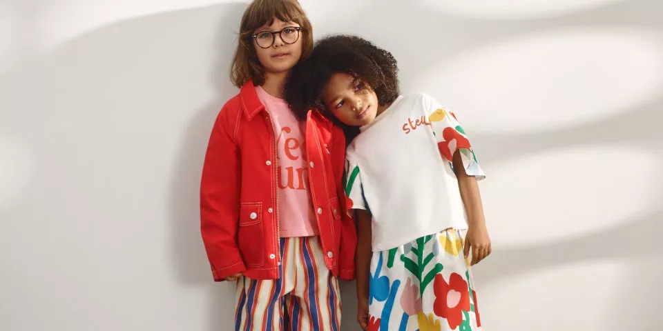 Two young girls in colourful kids’ outfits, one in red jacket and stripes, posing against a white studio wall