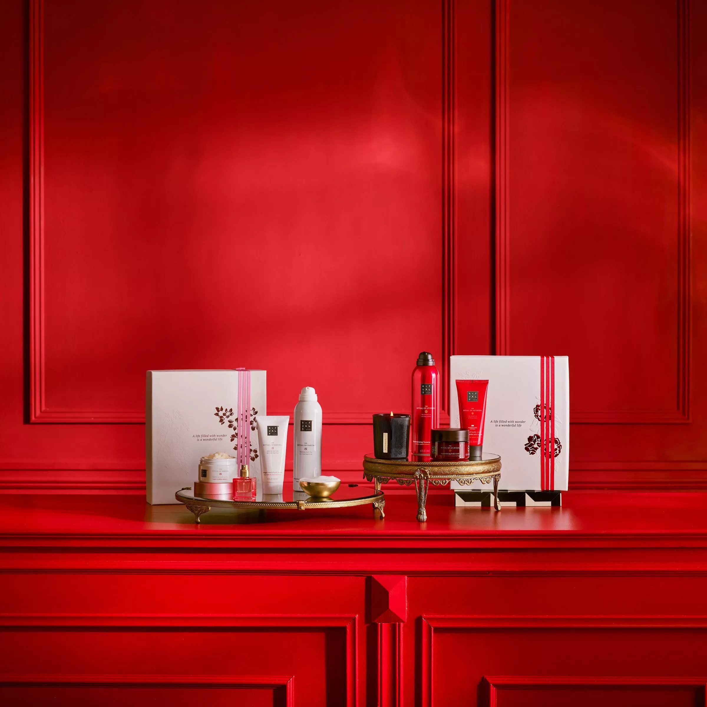 A luxury gift set by Rituals, featuring four white and pink packaged body care products (a scrub, a spray, a body cream tube, and a foaming shower gel) next to a large white and pink gift box, all sitting on a gold-rimmed tray against a red background