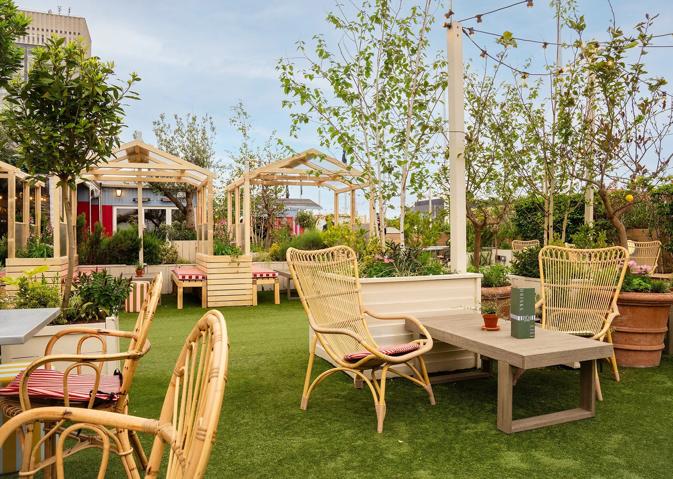 Outdoor seating area with wicker chairs, lush greenery, wooden structures, and vibrant plants under a clear sky.