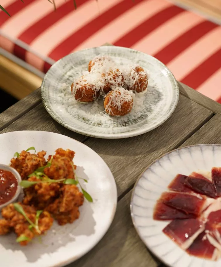Three plates of appetisers on a wooden table: arancini topped with cheese, spicy fried bites, and thinly sliced cured meat.