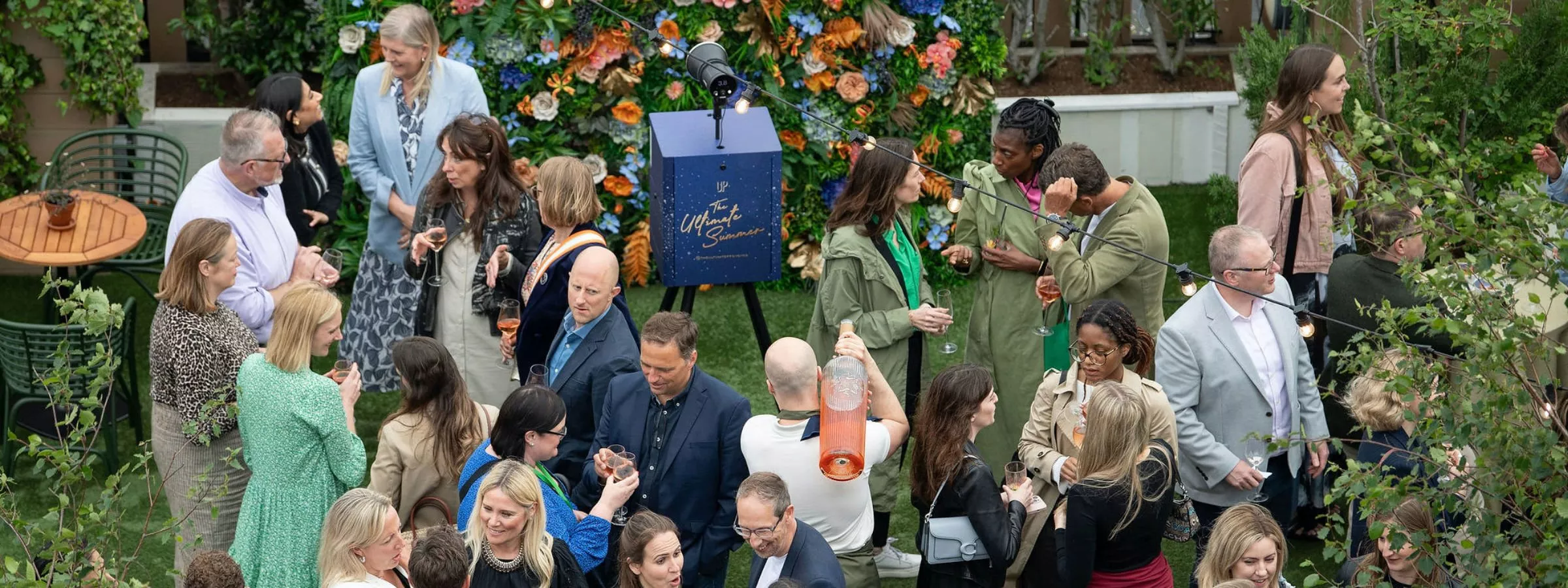 Aerial view of a lively garden party with guests mingling, holding drinks, and a vibrant floral backdrop.