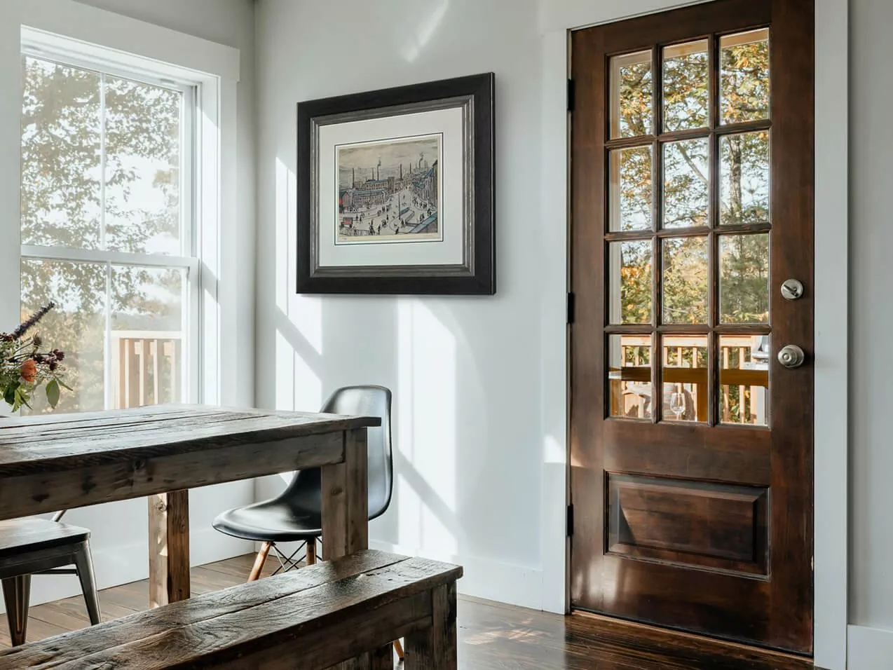 Sunlit dining nook with rustic wooden table, framed artwork, and glass-panelled door overlooking autumn trees
