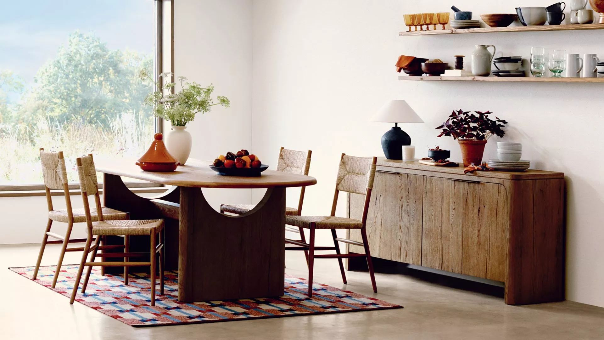 Modern dining room with wooden table and chairs, colourful rug, and shelves displaying crockery and plants.