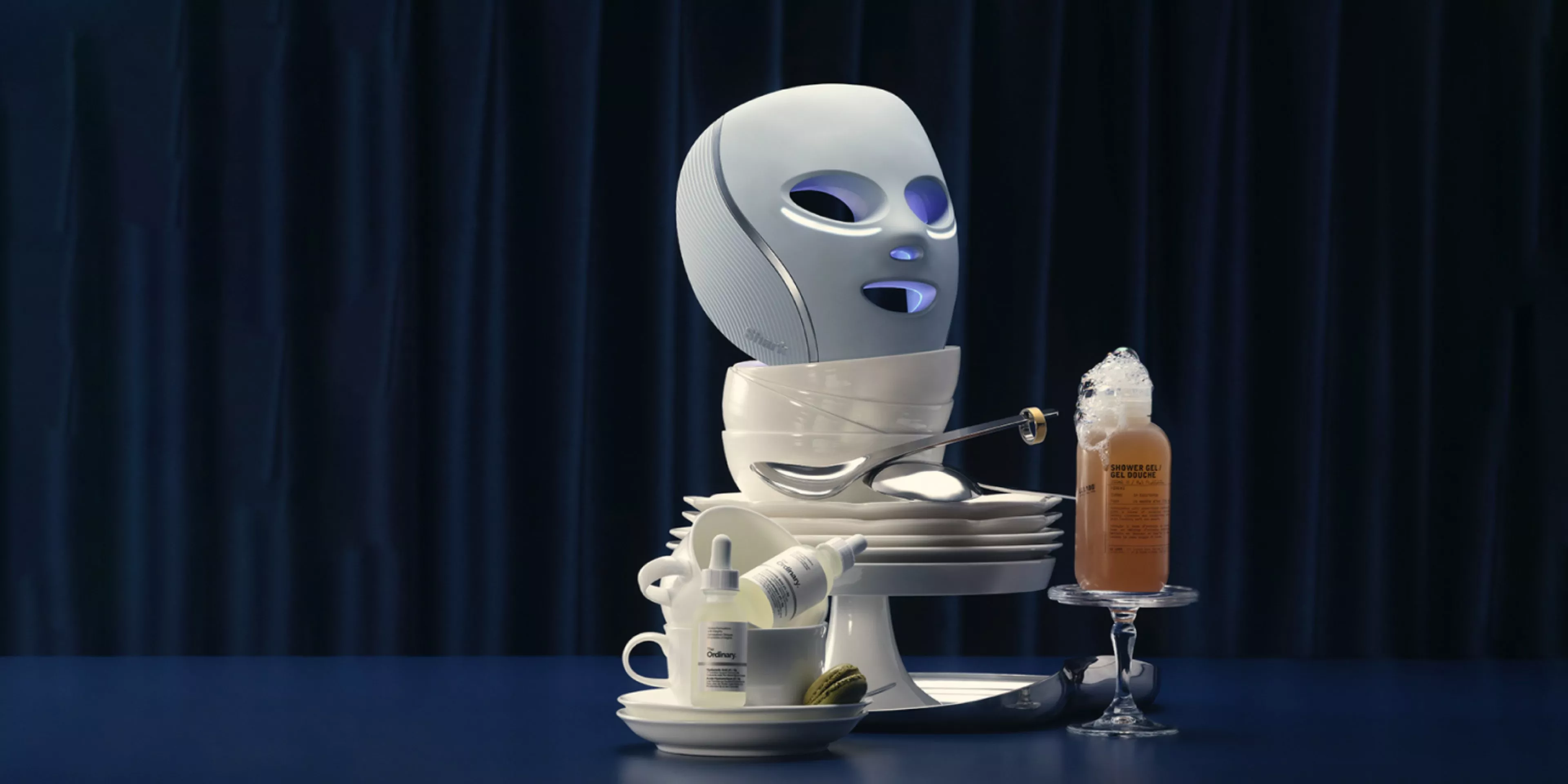 A futuristic white LED facial mask sits on a precarious stack of white porcelain bowls and plates, accompanied by a bottle of The Ordinary skincare and a tall glass of iced coffee.