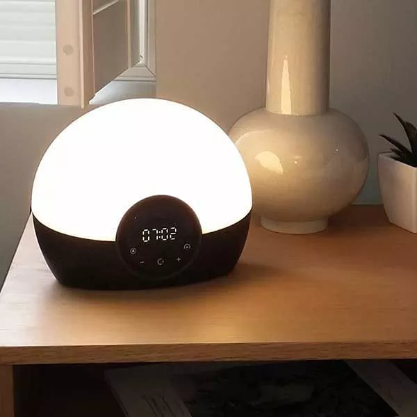 Modern sunrise alarm clock lamp with digital display on wooden bedside table, glowing warm white light