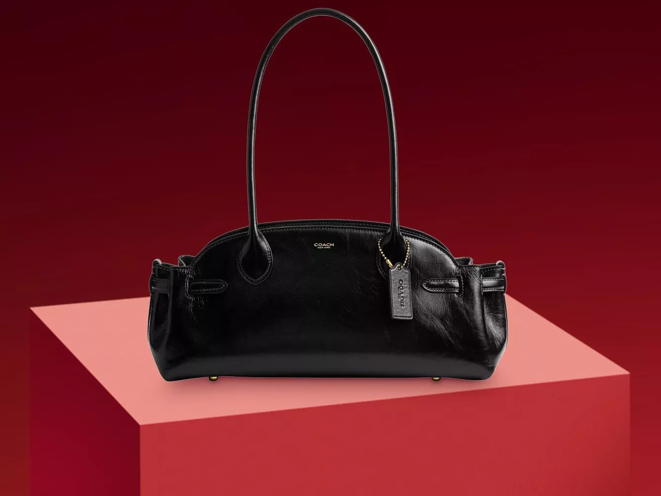 Black leather Coach shoulder handbag with long straps and hangtag, photographed on a red pedestal background