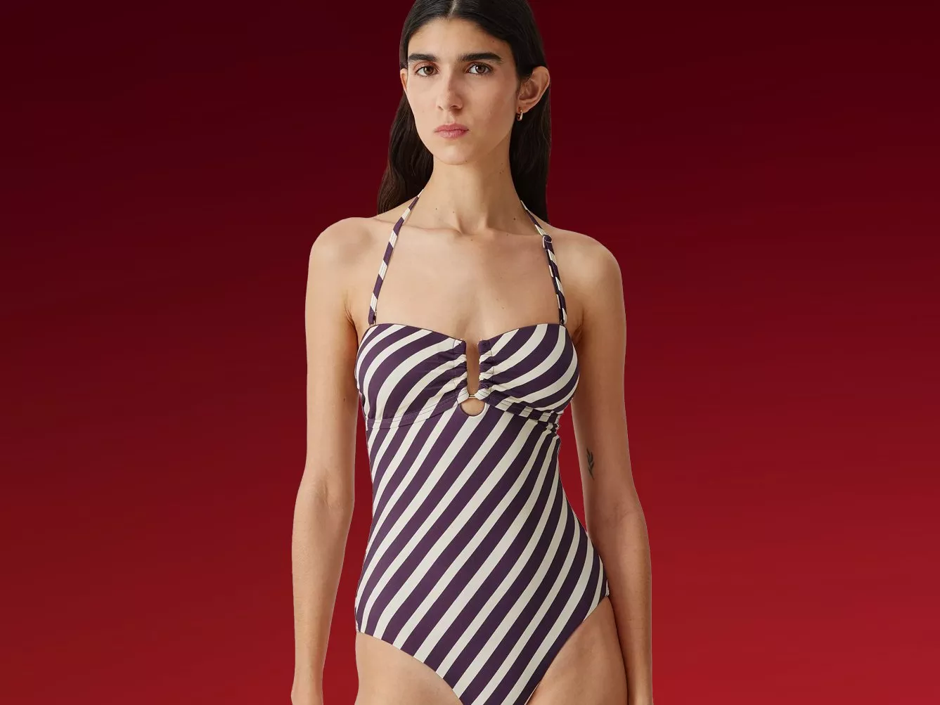 Model wearing a purple and white striped halterneck one-piece swimsuit, studio fashion photo on red background