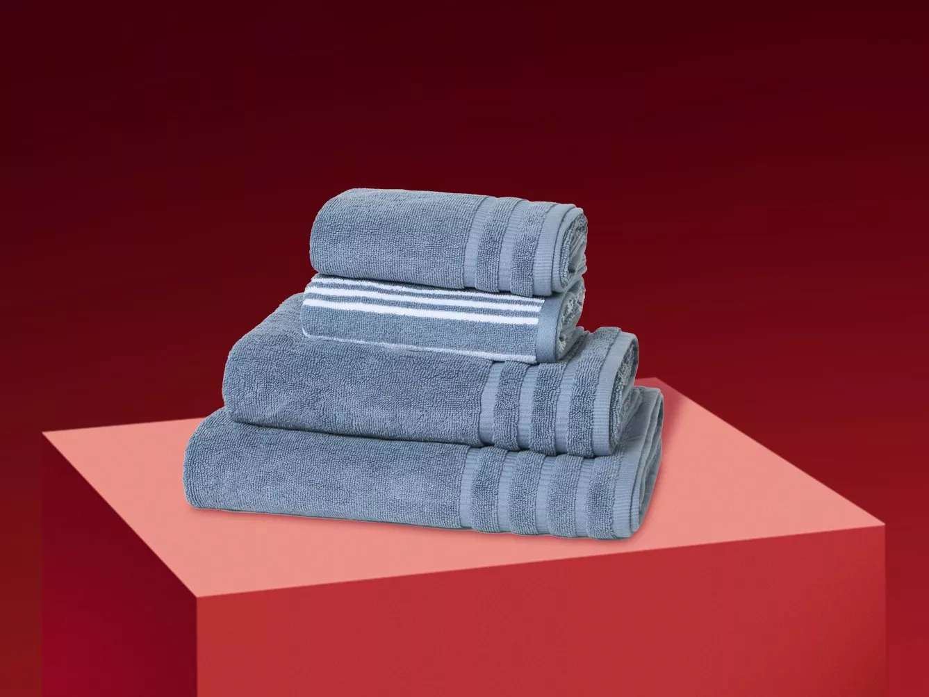 Stack of grey cotton bath towels with striped border on red pedestal, minimalist studio product photo