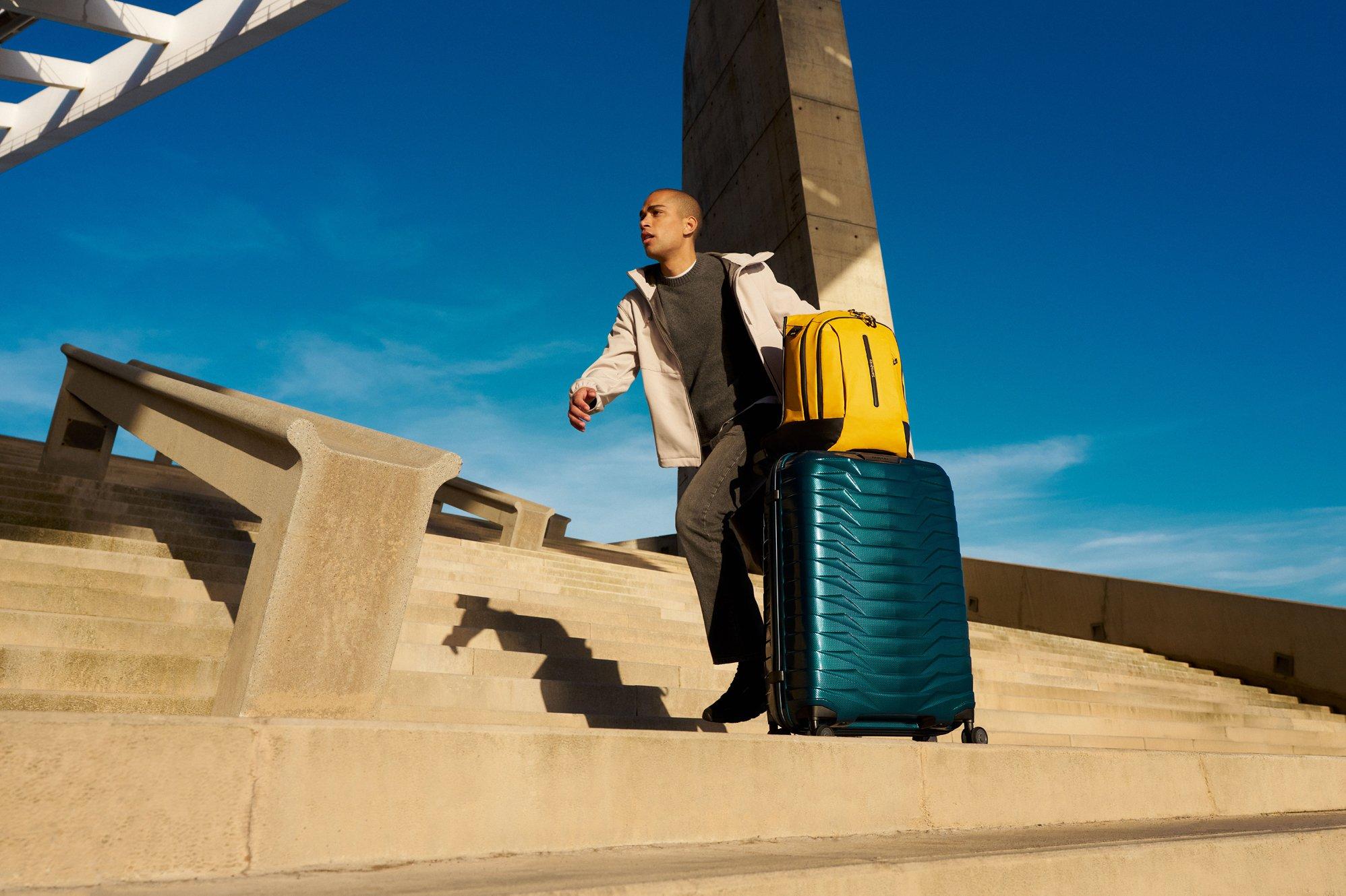 a man pushing a blue samsonite suitcase along a road in bright sunshine and blue skies