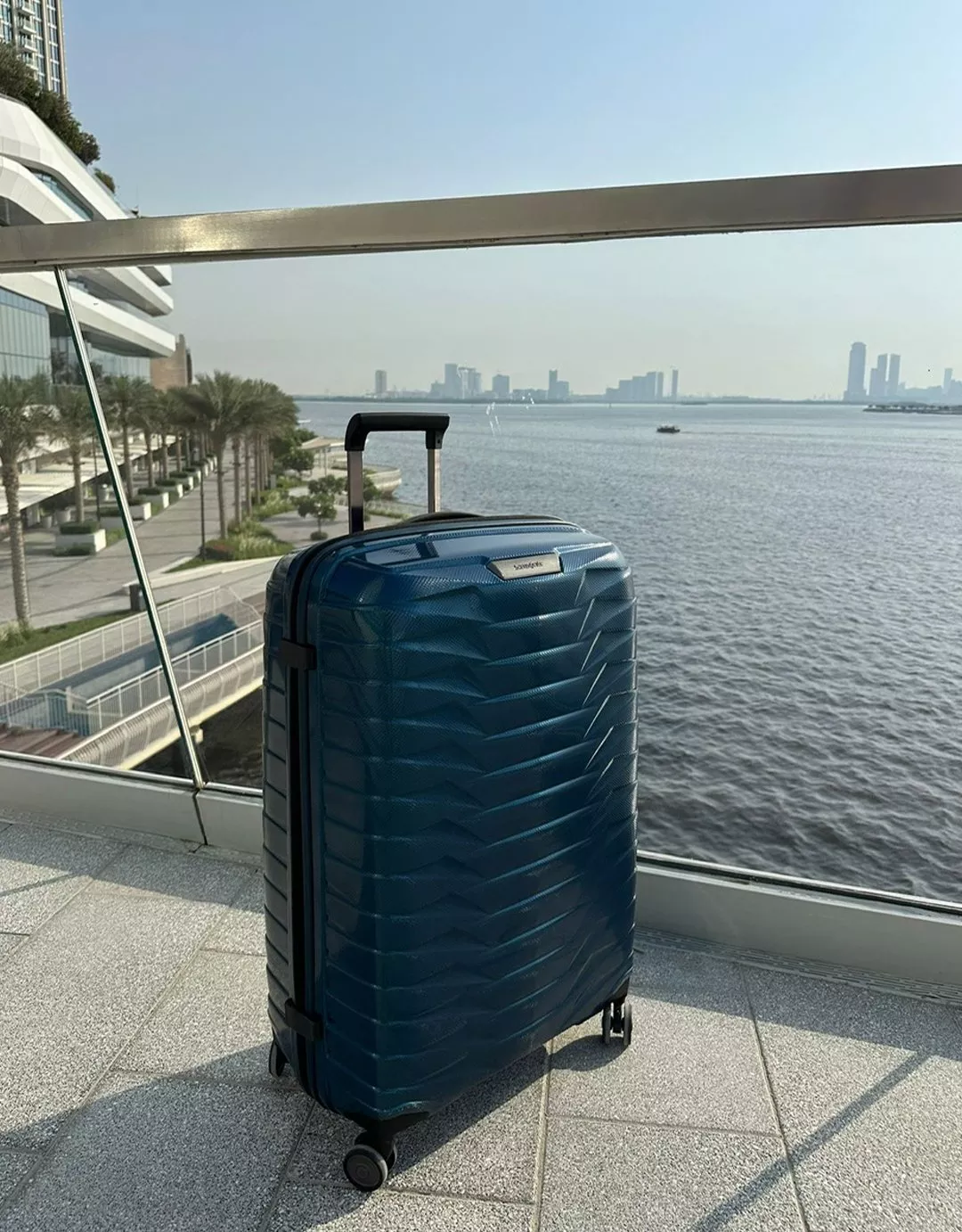 samsonite brand blue suitcase in front of a harbour with a city view in the background